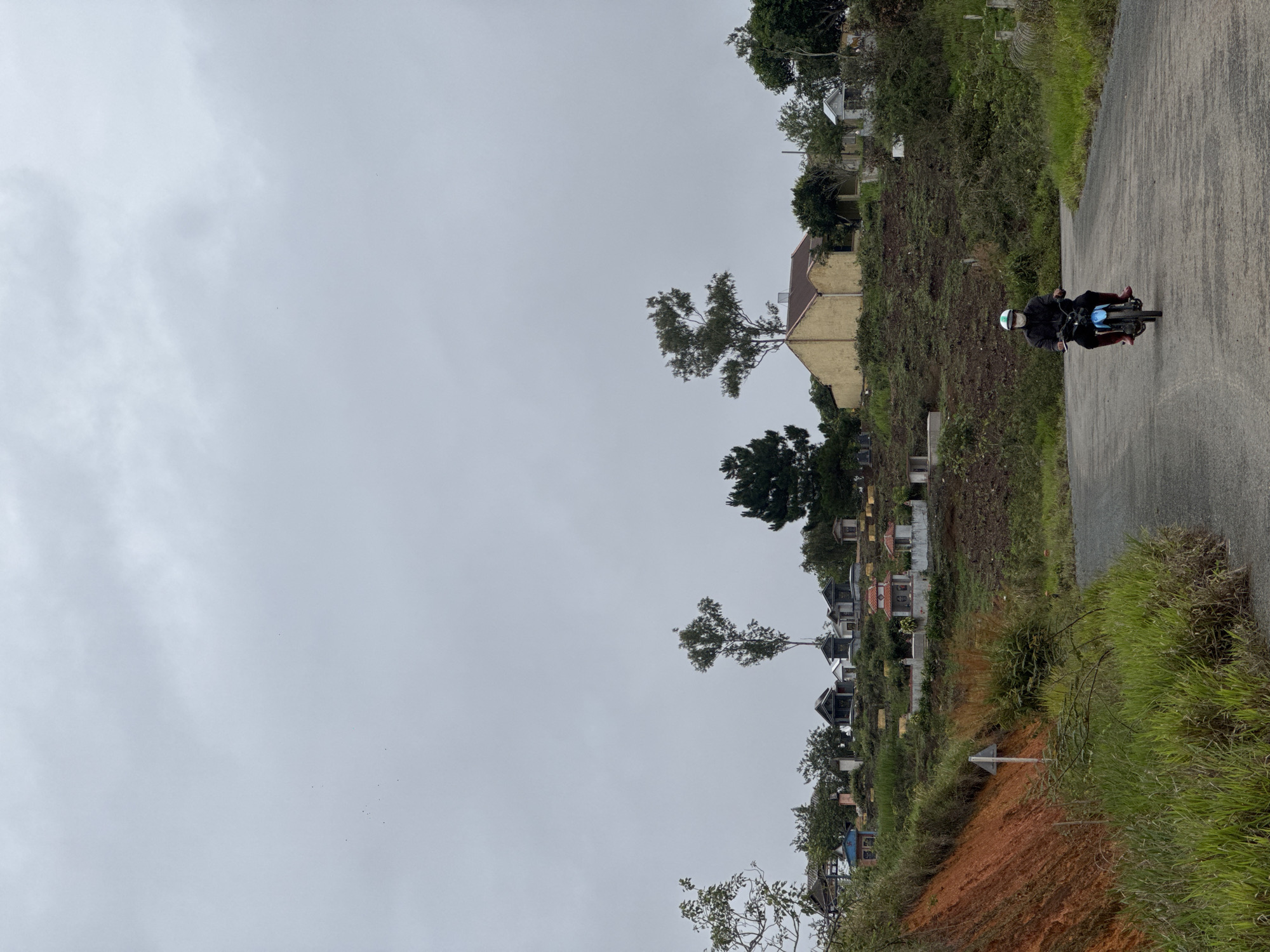 The scene depicts a motorcyclist traveling on a paved road in Trường Thọ, Vietnam. The rider wears a helmet and dark clothing, with bright red footwear. The road is lined with dense vegetation on the right and a terraced landscape with various structures and trees on the left. In the background are residential buildings and a predominantly overcast sky. The foreground includes a reddish-brown soil embankment.