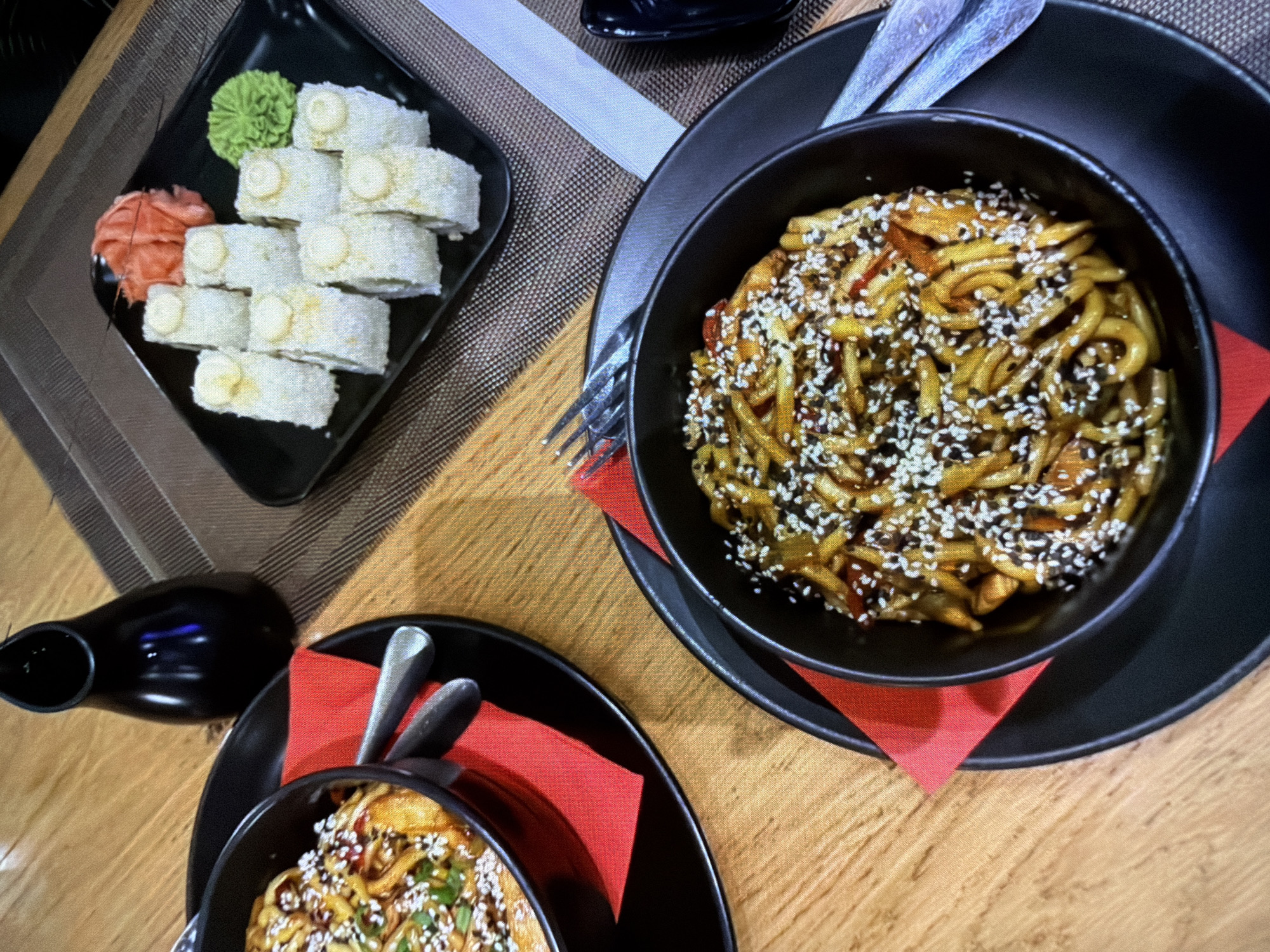 A top-down shot presents a sushi and noodle spread on a light brown wooden table in an unspecified location within the city of Unknown, Unknown. The main focus is a black bowl of noodles, generously sprinkled with sesame seeds and accompanied by pieces of what appears to be chicken and vegetables. This bowl sits on a square red napkin atop a larger black plate.
Adjacent to the noodle bowl is a black rectangular plate featuring a row of sushi rolls, served with wasabi and pickled ginger. To the side, a small black dish filled with soy sauce sits alongside a smaller bowl of noodles, complete with chopsticks resting on another red napkin. Cutlery is placed between the two noodle dishes, indicating preparation for consumption.