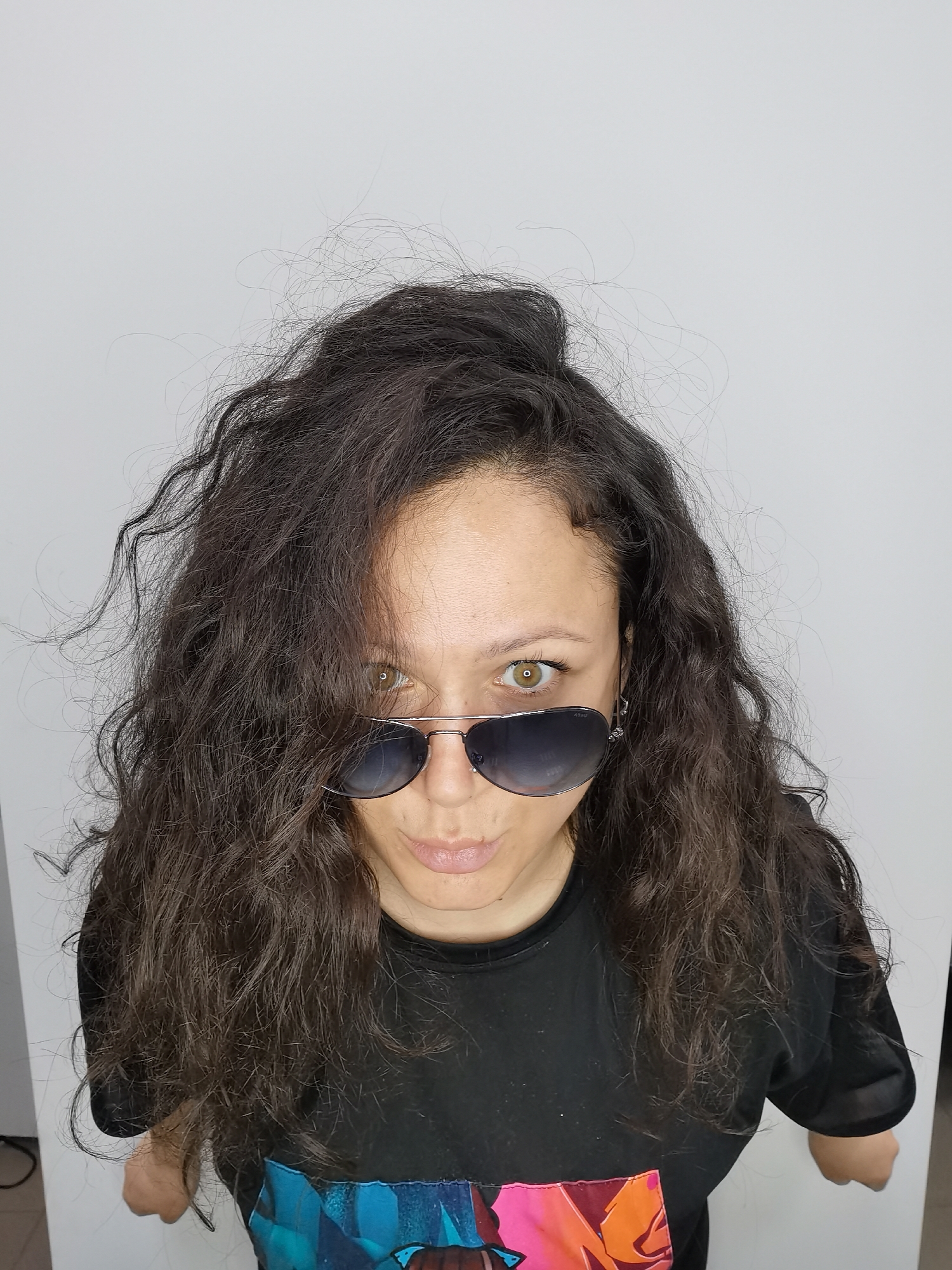 A woman with long, dark curly hair and olive skin is wearing aviator sunglasses and a black t-shirt. She appears to be indoors against a plain white wall. The lower portion of the shirt features colorful graphics. The location is not discernible from the image.