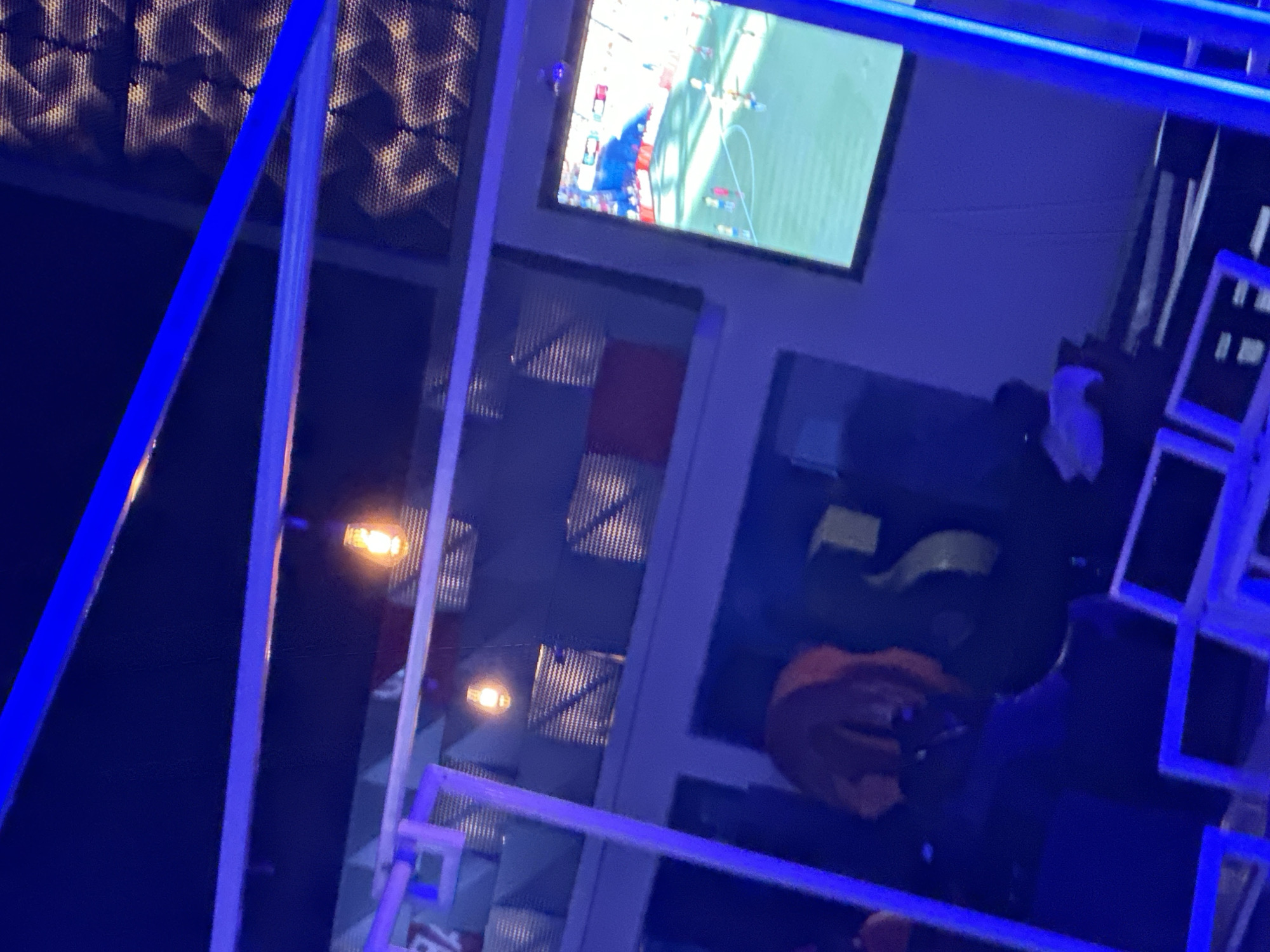 The image depicts an indoor scene, likely a bar or entertainment venue, with a dominant blue neon lighting scheme. In the background, a large flat-screen television displays a soccer match. The walls feature geometric textured panels. Several Edison-style light bulbs hang from the ceiling, emitting a warm orange glow. In the foreground, a person with orange hair is visible, wearing dark clothing. There are also glimpses of furniture and what appears to be a DJ booth. The overall ambiance is dark and dimly lit, consistent with a nighttime establishment. The location in Gura Top, Nigeria, is not directly discernible from the image content, but suggests an urban entertainment setting.