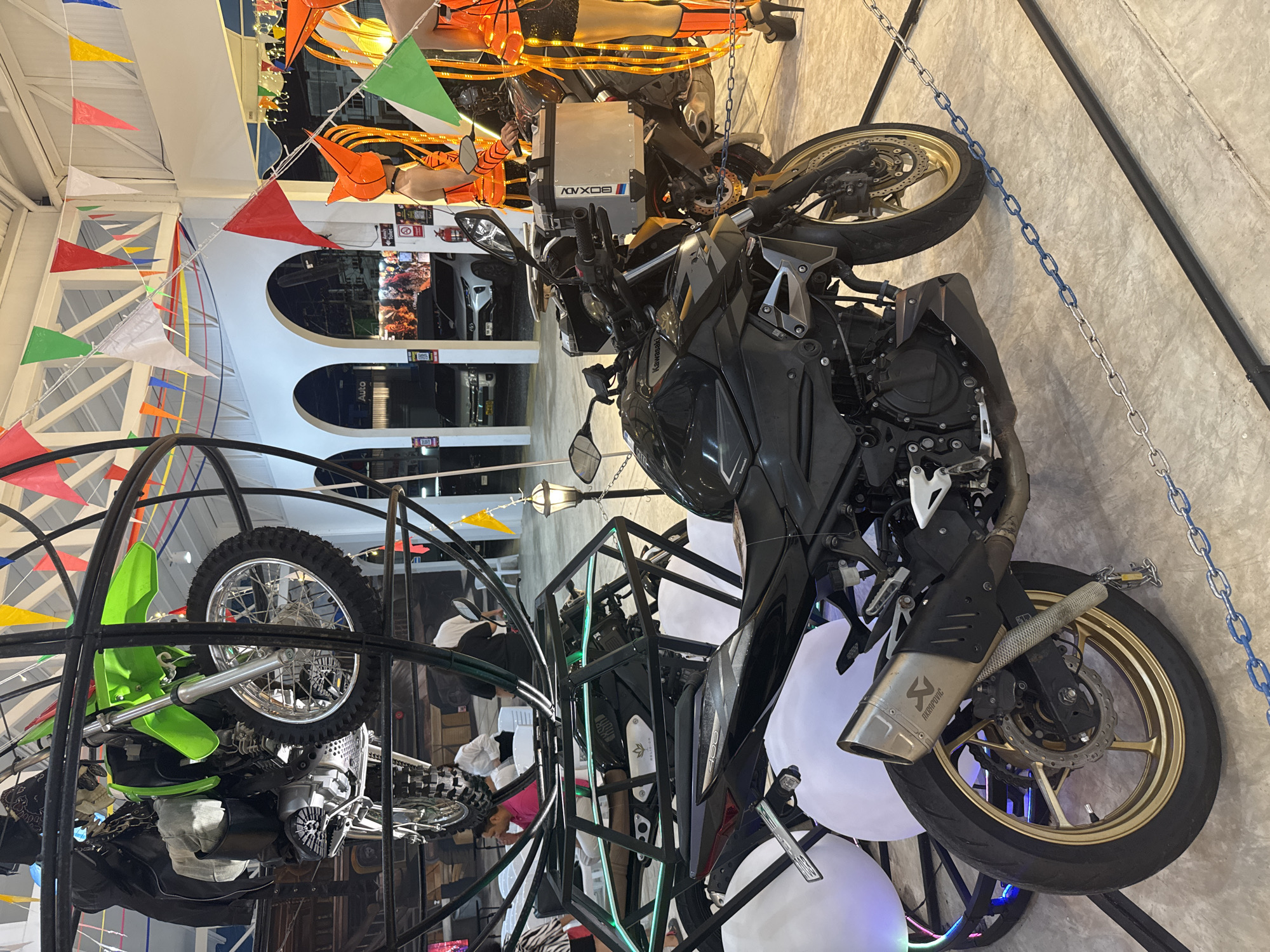 This is a photograph of a display featuring motorcycles, possibly in a showroom or at an event. The main subjects are two motorcycles, one primarily black and the other a bright green dirt bike. The black motorcycle is a sportbike with a gold-colored rear wheel and a distinctive Akrapovič exhaust. It is suspended by chains and positioned in the foreground, taking up a significant portion of the frame. The green dirt bike is housed within a circular black metal structure, its front wheel prominently displayed.
The setting appears to be indoors, with a concrete wall in the background and a white ceiling with structural elements visible. Decorative elements include strings of colorful triangular flags and orange lights, suggesting a festive atmosphere or a themed exhibition. Several white, balloon-like objects are also present, some illuminated with colored lights. In the background, there are glimpses of cars through arched openings, hinting at a larger venue.
There are no clearly identifiable people in the foreground, but silhouettes or partially obscured figures can be seen in the background behind the green dirt bike and within the arches. No specific activity is evident beyond the display of the motorcycles themselves. The lighting is artificial and varied, with some areas brighter than others, creating highlights and shadows. The overall impression is of a dynamic and visually engaging presentation of motorcycles.
Visible text includes "ADV 308" on a silver box on the black motorcycle and "AKRAPOVIĆ" on the exhaust pipe of the black motorcycle. The word "Kawasaki" is also visible on the fairing of the black motorcycle.