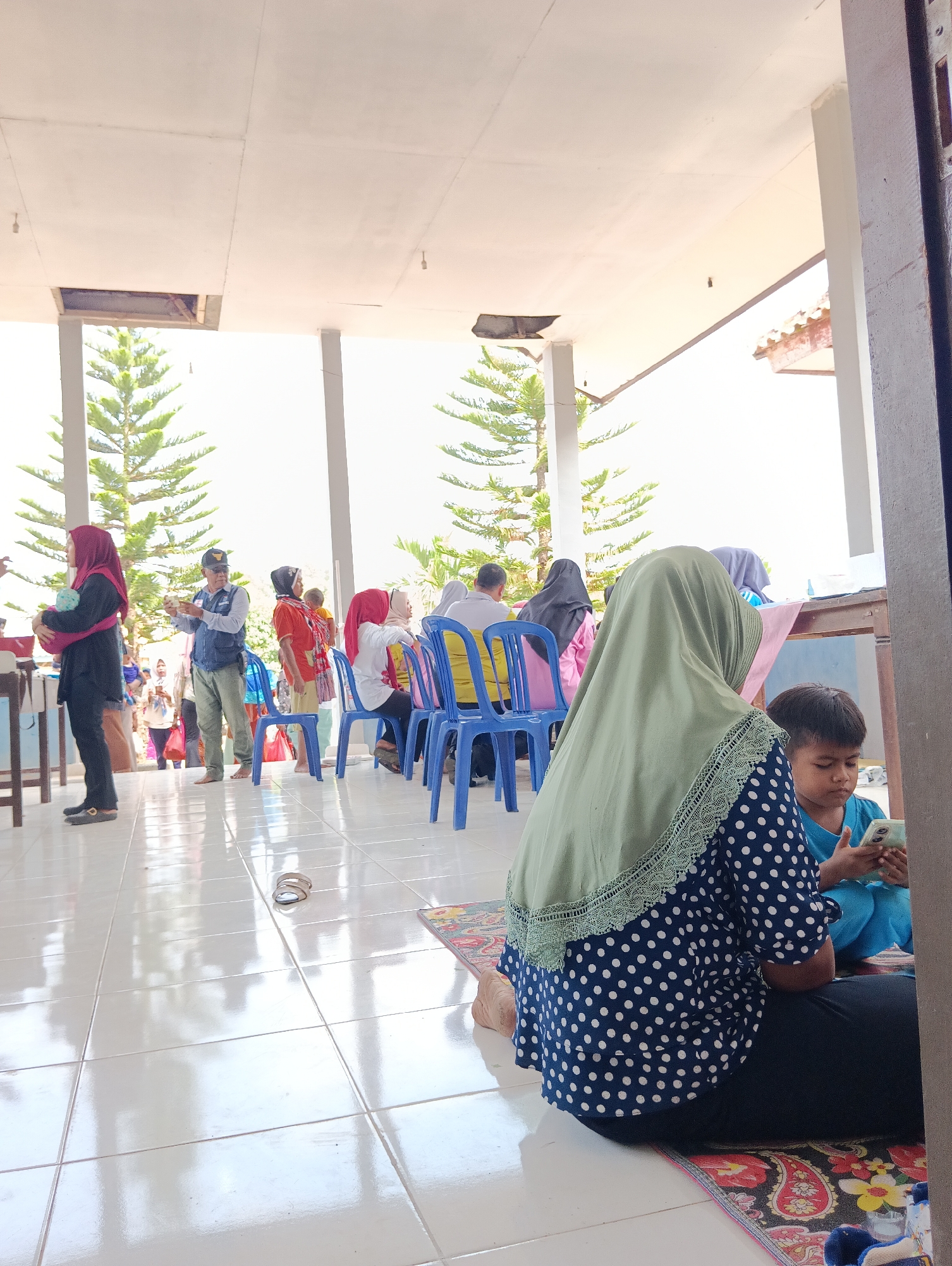 The image depicts an outdoor covered area, possibly a community hall or waiting area, with a tiled floor that reflects the bright natural light. In the foreground, a woman wearing a blue and white polka dot dress and a light green hijab is seated on the floor, facing away from the camera. She is looking down at a smartphone in her hands. To her right, a young boy in a blue shirt is also looking at a smartphone.
Behind them, several people are seated on blue plastic chairs, some facing forward and others with their backs to the camera. Among them is a woman in a red and white checkered scarf and orange top, and another woman in a pink shirt and black hijab. A man in a blue vest and a cap stands in the middle ground, holding what appears to be money and a paper. To the left, a woman in a dark outfit and red hijab is holding a baby.
The background shows tall, slender evergreen trees and a bright, overcast sky. The architecture of the building includes white pillars supporting a light-colored ceiling. A table with some items is visible in the upper right portion of the image. The overall impression is of a casual gathering or waiting area, potentially for an event or distribution. The image is shot from a low angle, emphasizing the tiled floor and the seated figures.