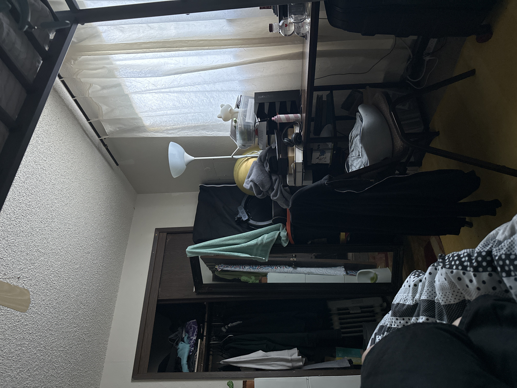 The image depicts an indoor scene, likely a bedroom, with the camera positioned at an angle that makes the content appear rotated. The room is cluttered with various objects, including clothing draped over furniture and shelves, a desk with items on it, and a wardrobe.
Dominating the scene is a wardrobe with wooden doors and shelves. The shelves are filled with folded clothes, towels, and other miscellaneous items. A mint green towel is prominently displayed on an upper shelf. Below, the shelves hold various garments, including patterned scarves and dark-colored clothing.
To the left of the wardrobe, a white floor lamp with a conical shade stands near a textured, light-colored wall. In the background, sheer white curtains hang, allowing diffused light to enter the room, suggesting it is daytime.
A desk is situated behind the lamp, with several items on its surface, including what appear to be bottles, a small white bear figurine, and headphones. A laptop or computer monitor is also visible on the desk.
Further to the right, a black suitcase is partially visible, suggesting travel or storage. The floor is covered in a yellow-toned material. In the foreground, a person is partially visible, dressed in black, and lying on a bed with a black and white patterned comforter.
The overall impression is one of a lived-in, somewhat untidy personal space. The lighting suggests it is daytime. No specific time of day or weather can be determined, nor can any specific emotions be inferred. There is no visible text in the image. The location context provided is Vancouver, United States of America, but this cannot be confirmed from the image alone.