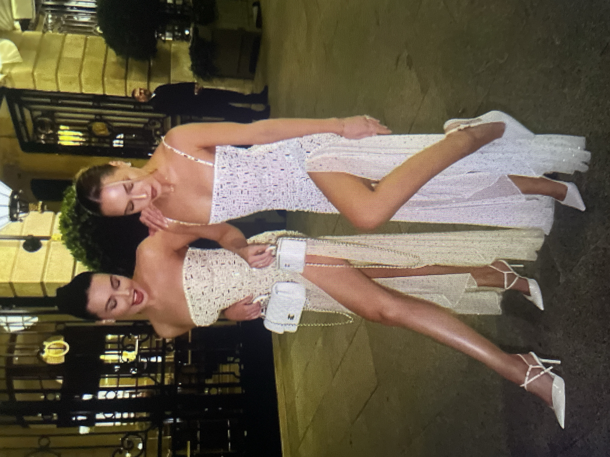 Two women, dressed in glamorous, sparkly evening gowns with high slits, pose outdoors at night in front of a building with ornate architecture. The woman on the left wears a strapless gown and holds a small white handbag. The woman on the right wears a spaghetti strap gown and also holds a small white handbag. Both women are wearing white, pointed-toe high heels. The ground is paved and appears wet, reflecting the ambient light. A man in a dark suit stands in the background, appearing to be a security guard or attendant. The overall setting suggests a formal event or a night out in a city. The image does not contain any elements that would allow it to be identified as being in Kołobrzeg, Poland.