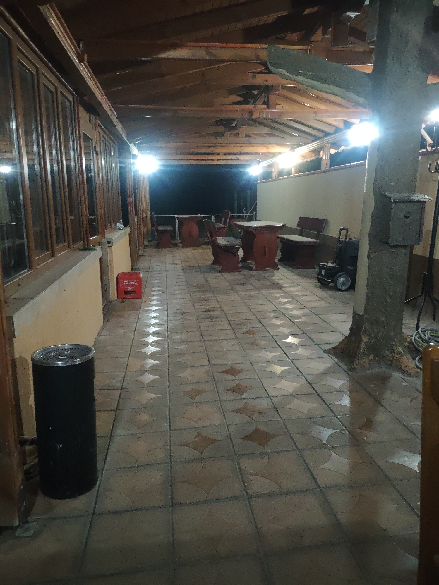 The image captures a semi-enclosed patio or veranda at nighttime, featuring a wooden ceiling structure with exposed beams. The flooring is tiled with a decorative pattern of overlapping circles that create diamond shapes. Numerous bright overhead lights illuminate the space, casting strong reflections on the seemingly wet floor.
On the left side, a long wall with multiple windows, framed in brown wood, extends into the distance. Below the windows, there's a light-colored ledge or low wall. In the foreground on this side, a black cylindrical trash can with a metallic lid is visible. Further along the wall, a red plastic crate, with the Cyrillic text "Минерална вода" (Mineral water) partially visible, sits on the ground.
Towards the right, a thick, rough-textured pillar, resembling a tree trunk, stands prominently. A small, grey box is attached to this pillar, and several hooks are visible on the far right. Behind the pillar, a black utility cart or speaker on wheels is parked. Coiled hoses are also present near the base of the pillar.
In the mid-ground and background, several dark red wooden tables and benches or chairs are arranged for seating, suggesting a dining or lounging area. The background beyond the patio is dark, indicating it is night. No people are visible in the scene, which appears quiet and unoccupied.
