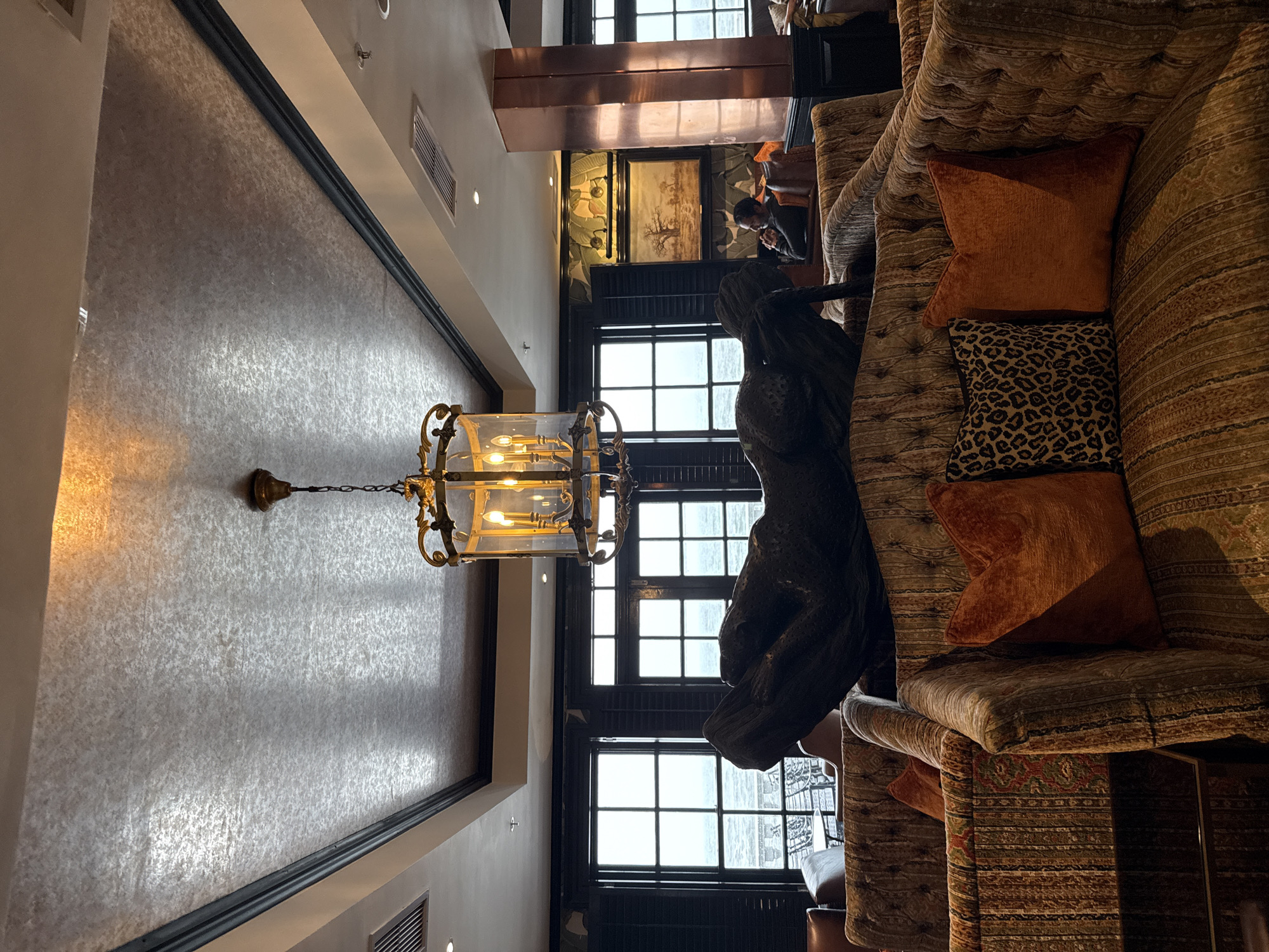 Here is a description of the image:
The image depicts an interior setting with a blend of classical and contemporary elements. The scene appears to be a lounge area or a lobby of a building, possibly a hotel or a high-end residential complex.
A prominent feature is a large, ornate chandelier suspended from the ceiling. It is a cage-like structure enclosing light bulbs, giving off a warm, inviting glow. Adjacent to the chandelier is a textured wall that creates a visually interesting background.
To the right, there is a plush, patterned sofa adorned with decorative pillows in varying textures and colors, including a leopard print. A dark, abstract sculpture stands near the sofa, adding an artistic touch to the space.
Large windows with black frames are visible, offering a view to the outside and letting in natural light. The windows, along with the lighting fixtures, suggest that this space is located in an urban area, though the specific city and neighborhood are not identifiable.
In the background, a person can be seen sitting at a table, possibly engaged in conversation or working. Artwork adorns the walls, contributing to the sophisticated ambiance of the place.