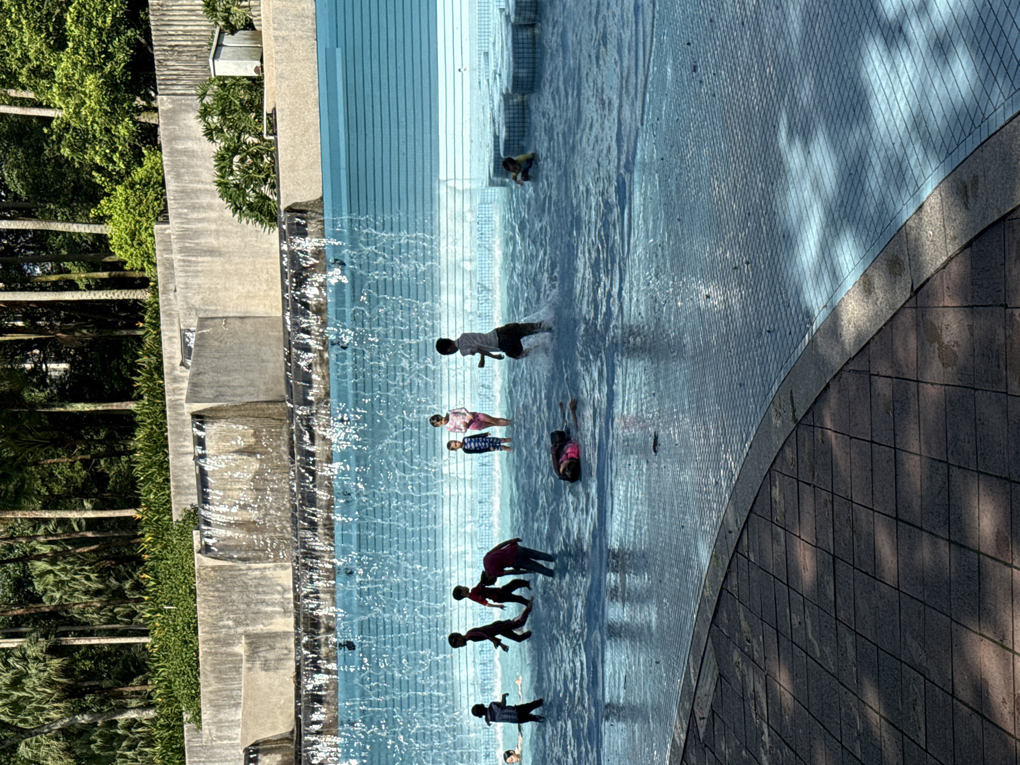 The image depicts a water feature with multiple individuals, likely children, interacting within the water. The location appears to be a public space in Kuala Lumpur, Malaysia. The water feature is characterized by stepped tiers with water cascading down. The surrounding environment includes trees, greenery, and stone or concrete structures. Some of the children are wading or running through the water, while others appear to be sitting or playing near the edges. The ground surrounding the water feature is paved.