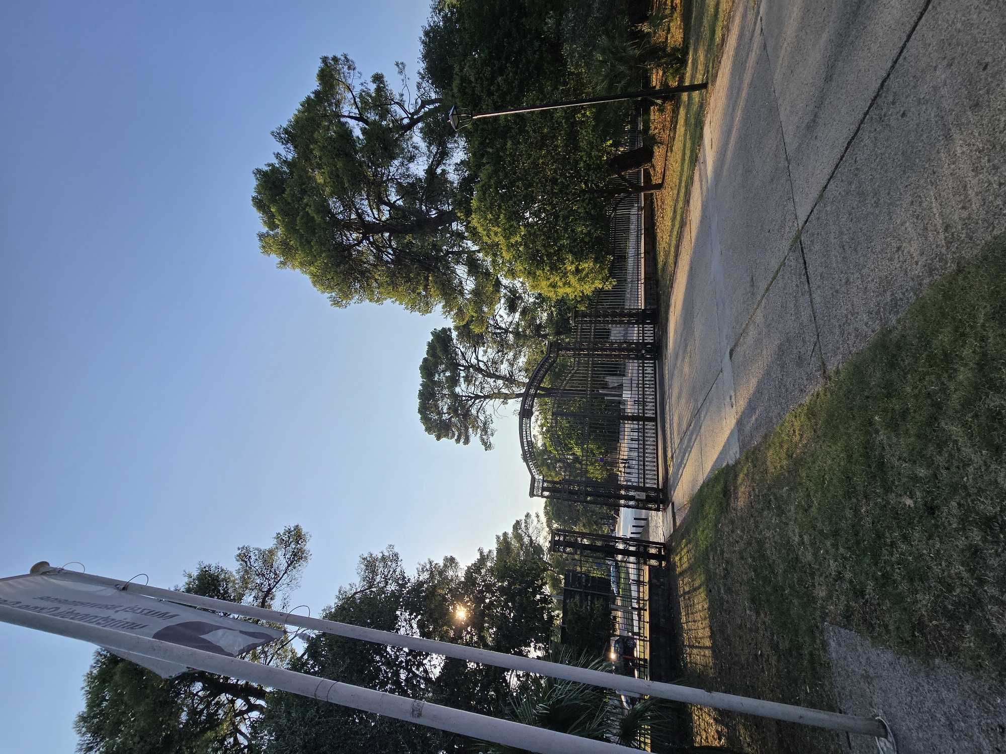 A wide-angle, low-angle shot captures the entrance to a park in Podgorica, Montenegro. A tall flagpole stands to the left, bearing a white flag with dark text and an image. Sunlight filters through the leaves of lush green trees, casting dappled shadows on a paved walkway leading towards a large, ornate black metal gate. The gate is partially open, revealing more trees and a lamppost beyond. The sky is a clear, bright blue, with the sun positioned high and slightly to the left, indicating it is likely late afternoon or early morning. The grass on the left side of the walkway appears dry and sparse in places, contrasting with the vibrant green of the surrounding foliage. A few cars are visible through the gate, suggesting the park is accessible by road. The overall impression is one of a peaceful, well-maintained park entrance bathed in warm sunlight.