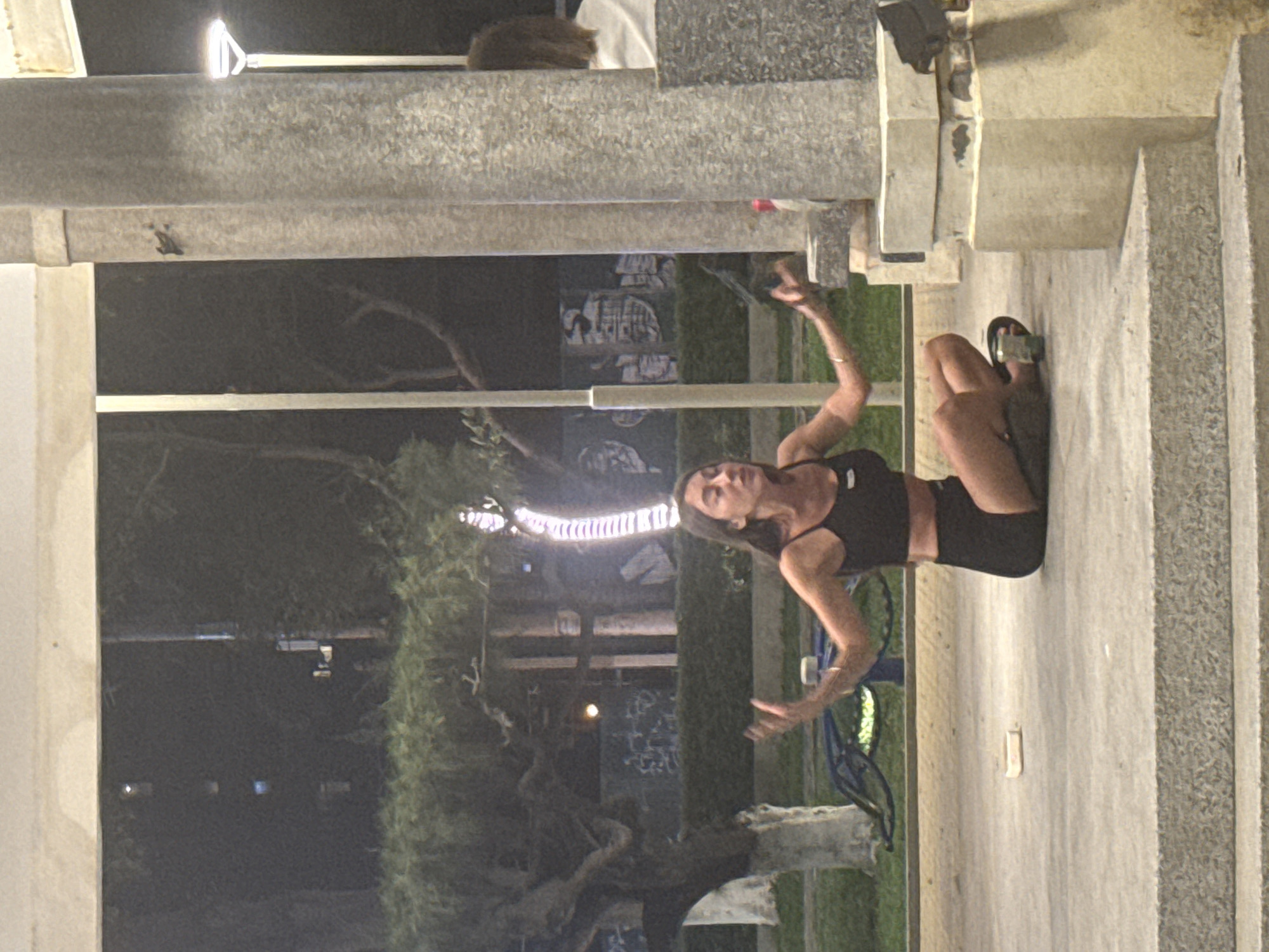 A woman in athletic wear stands in an outdoor area of Nha Trang Ward, Vietnam. She has her arms raised, gesturing as if performing a yoga pose or dance. She is positioned on a concrete surface adjacent to what appears to be a park or green space with trees and some urban furniture like a blue metal structure that looks like exercise equipment. Overhead, there are decorative lights strung among the trees. A large, grey concrete structure forms the upper border of the frame, possibly part of a building or an architectural feature of the area. The time appears to be evening or night.