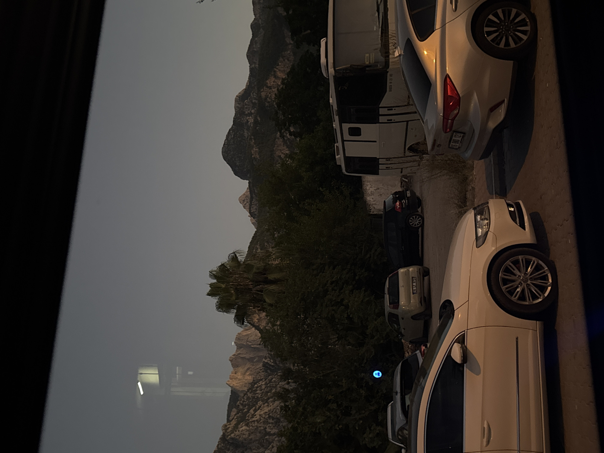 The image shows a view from inside a vehicle, looking out at a parking lot with several cars and a bus. In the background, there are lush green trees and rocky mountains. The sky is a dusky gray, suggesting either dawn or dusk. The scene appears to be in the city of Kemer, Turkey, given the distinctive mountainous terrain and palm trees. On the left, a white sedan is parked, its front facing left. To its right, a silver sedan is parked, facing right, with its taillights illuminated. A white bus is parked further back, with a white roof-mounted air conditioning unit visible. Several other cars are parked in the lot, including a light-colored compact car and a dark-colored compact car. A blue light is visible in the distance among the trees. The foreground shows the interior of a vehicle, with a dark frame at the top of the image.