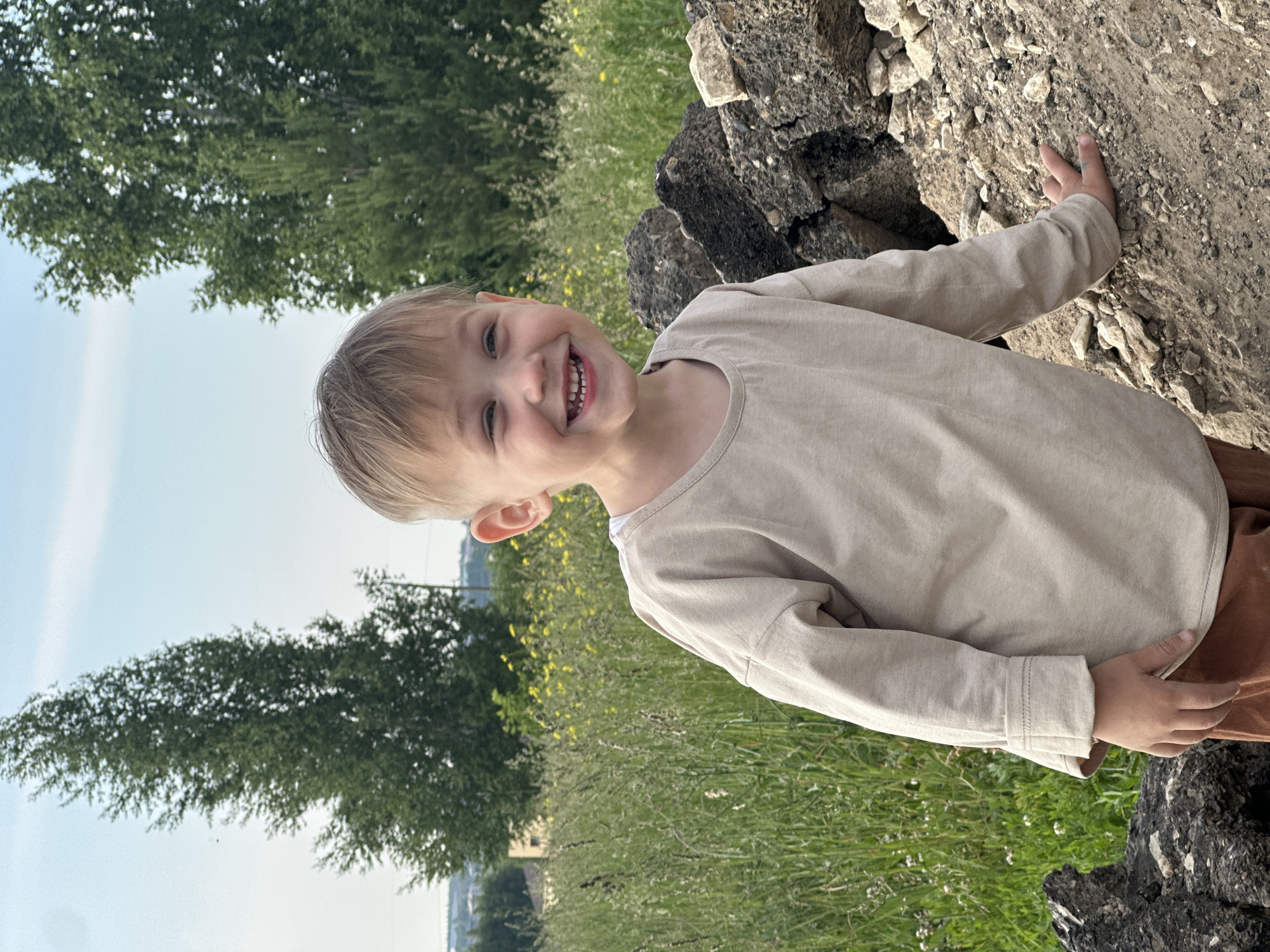 A young boy with blond hair is smiling and looking to his left in front of a pile of rocks and a grassy field. He is wearing a light beige long-sleeved shirt and brown pants. In the background, there are tall green trees and a clear sky. The location is the city of Estachi, Russia.
