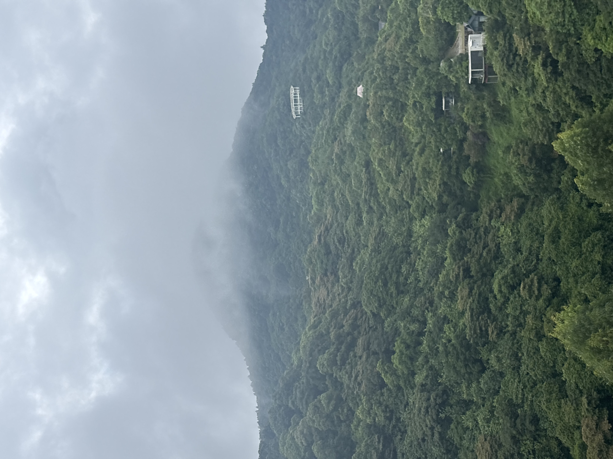 This image depicts a view of a misty mountain landscape with lush green foliage and several buildings nestled into the hillside. The mountain peak in the background is partially obscured by clouds, creating a soft, atmospheric effect. In the middle ground, a distinctive circular structure with white supports is visible on the slope. Further down the hillside, several modern buildings with clean lines and balconies are scattered amongst the trees. One building in the foreground appears to be a multi-story house with a large balcony. The environment is clearly a natural, mountainous area with dense vegetation. The image is not specific to Guangzhou City, China, as no identifying landmarks or contextual elements suggest this location.