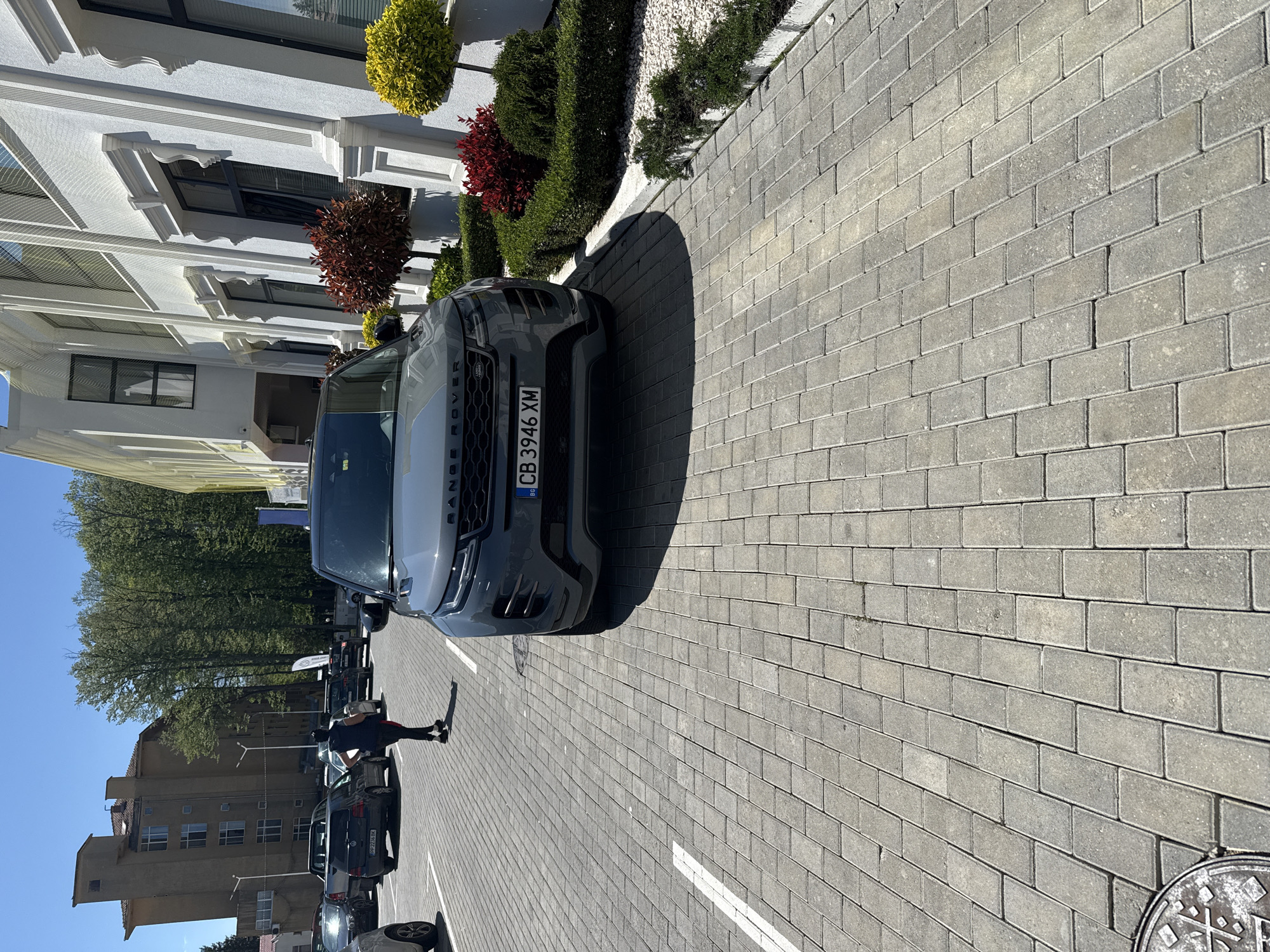 The image shows a gray Range Rover parked on a gray brick-paved street, with the license plate "CB 3946 XM" visible. The vehicle is positioned near a building with white facade and decorative elements, accented by landscaping including shrubs and trees.
Further down the street, a person is walking past parked cars. Additional buildings are visible in the distance. The location is Unknown, Unknown.