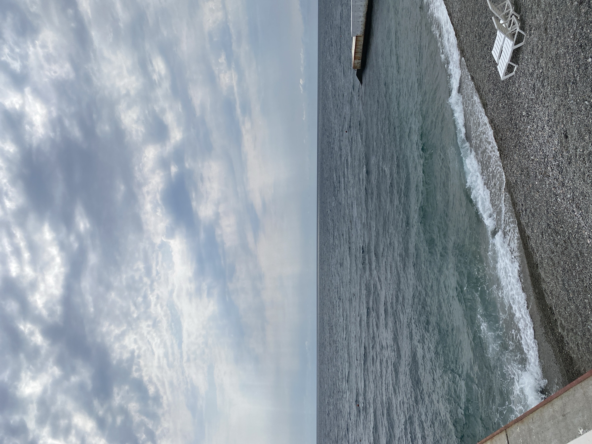 The image depicts a view of the Black Sea coast in Gasprinskyi settlement council, Crimea, Ukraine. The sky is overcast with grey clouds, and rays of light are breaking through in the upper left portion of the frame. The sea is a deep blue-grey with gentle waves lapping onto a pebbled beach. On the right side of the image, a concrete pier extends into the water. Two white lounge chairs are positioned on the pebbled beach in the foreground. The overall scene conveys a sense of calm and overcast weather.