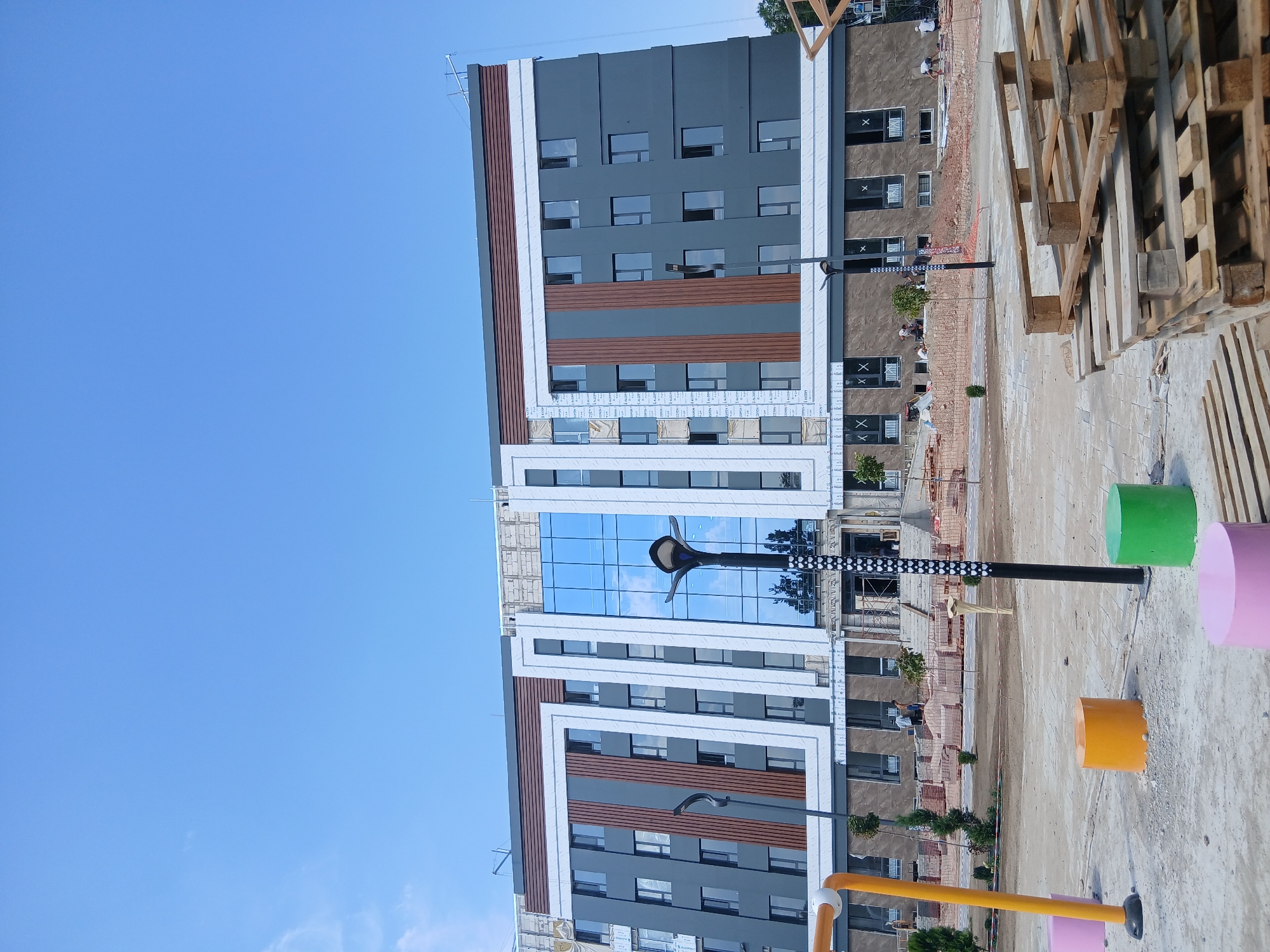 This is a wide, low-angle, eye-level outdoor shot of a modern building under construction in the city of Talas, Kyrgyzstan. The building is multi-story, with a dark gray facade and distinctive brown and white vertical accents. A large central section features a wall of blue glass windows, reflecting the clear blue sky. Several floors of the building have rows of dark-framed windows. Construction materials, including stacks of wooden pallets and what appear to be bricks, are visible in the foreground and around the building's base. In front of the building, there are colorful cylindrical seating elements in pink, orange, and green, as well as a yellow playground structure. Several individuals, dressed in casual attire, are scattered around the construction site, some appearing to be workers. A modern black lamppost with a distinctive top stands in the middle ground. The overall scene suggests an active construction site in an urban or developing area.