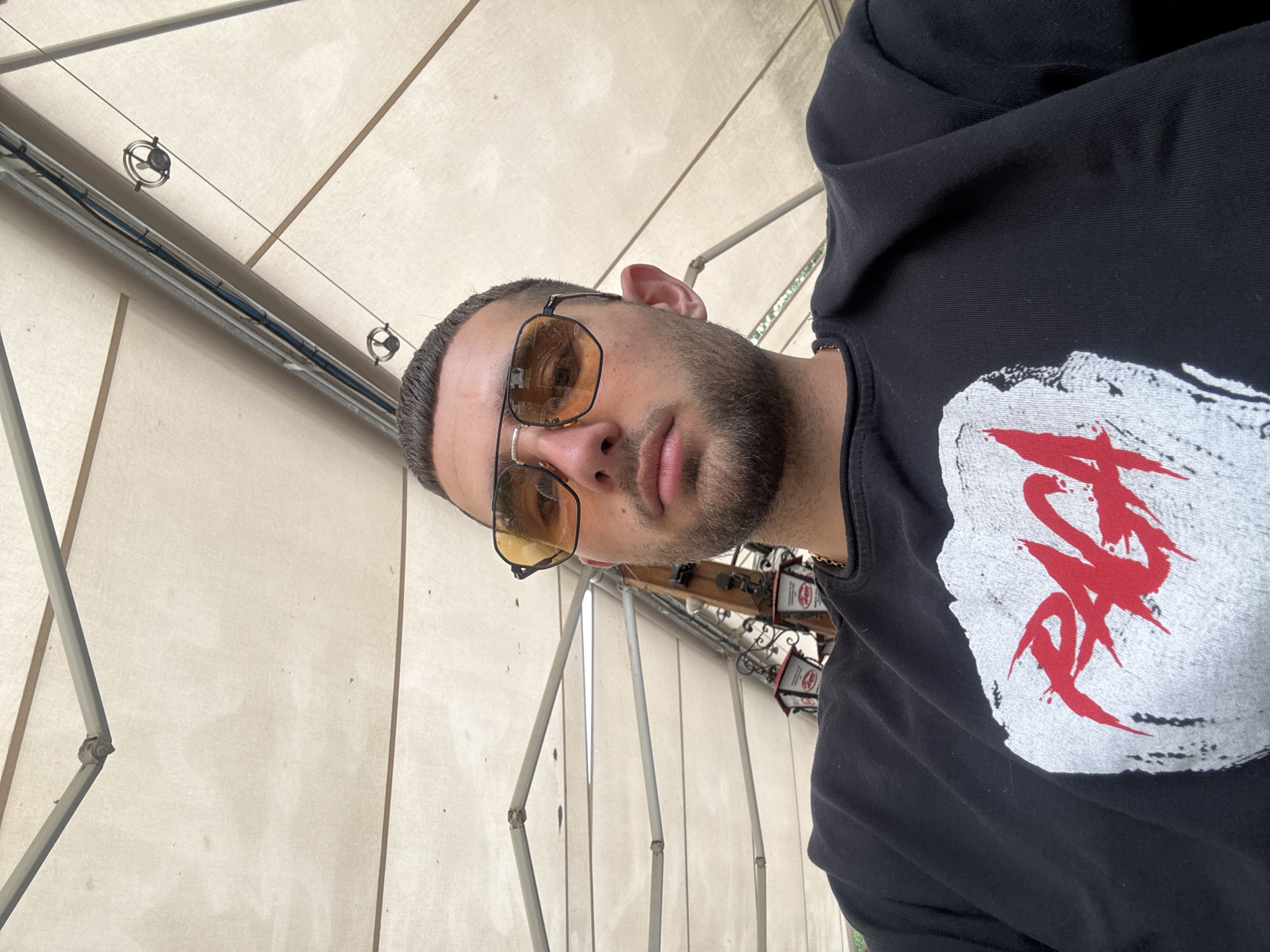 A close-up, tilted portrait shot of a man wearing sunglasses and a black sweatshirt with a white and red design. The background suggests an outdoor café setting. A cream-colored awning and metal framework are visible. The café might be located in Ruse, Bulgaria but there is no information in the image to support or refute this statement.