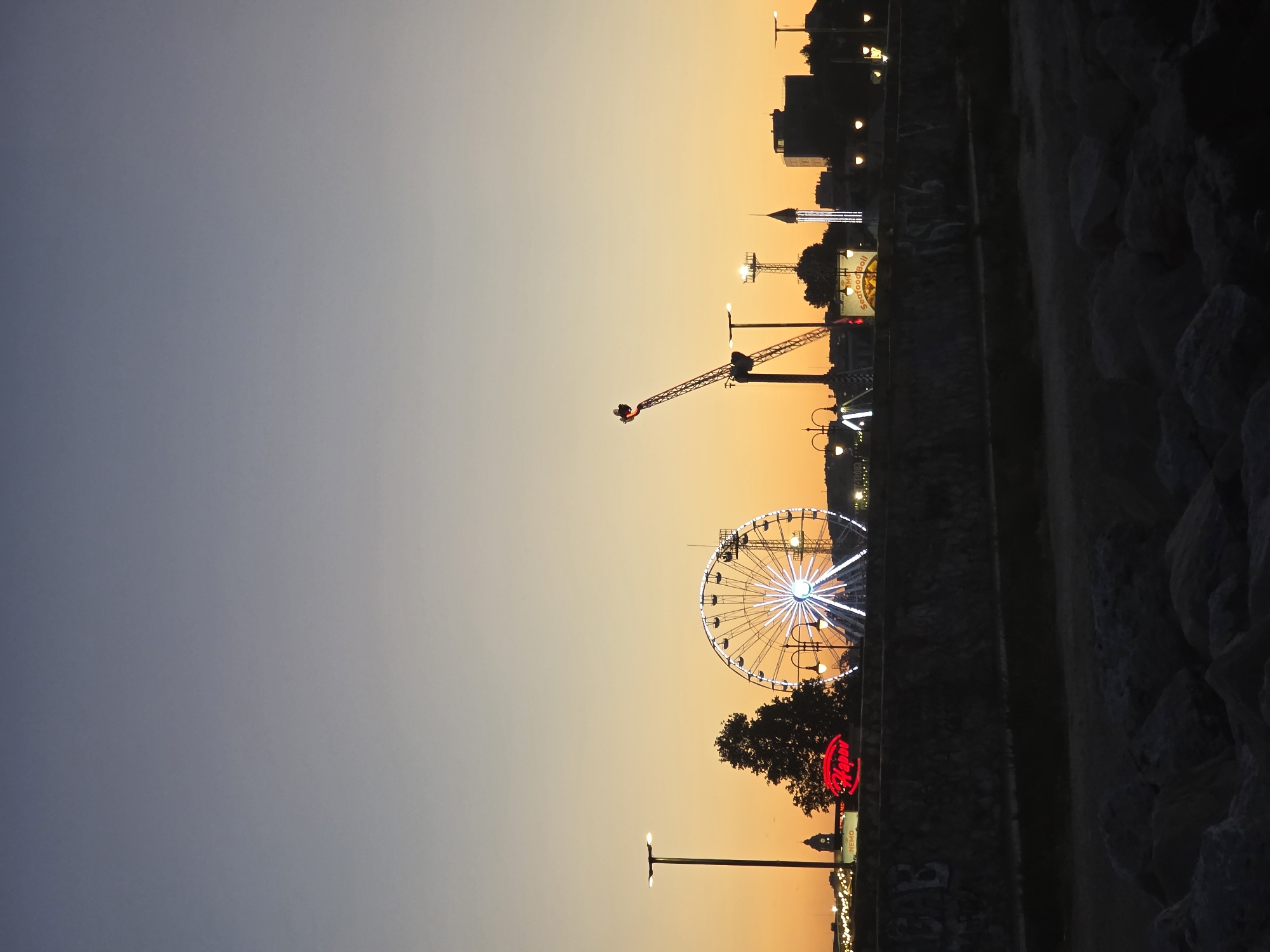 The image captures a waterfront amusement park in Varna, Bulgaria, during twilight. A Ferris wheel, illuminated with white lights, dominates the center-left of the frame. To its left, a red neon sign reads "Happy". A tall amusement ride, a swing ride with a rider at the top, is visible on the right. Silhouetted against the orange and purple sunset sky are various buildings and streetlights. In the foreground, large rocks line a dark, shadowed area, likely the shoreline. The overall scene depicts a lively entertainment area at the end of the day.