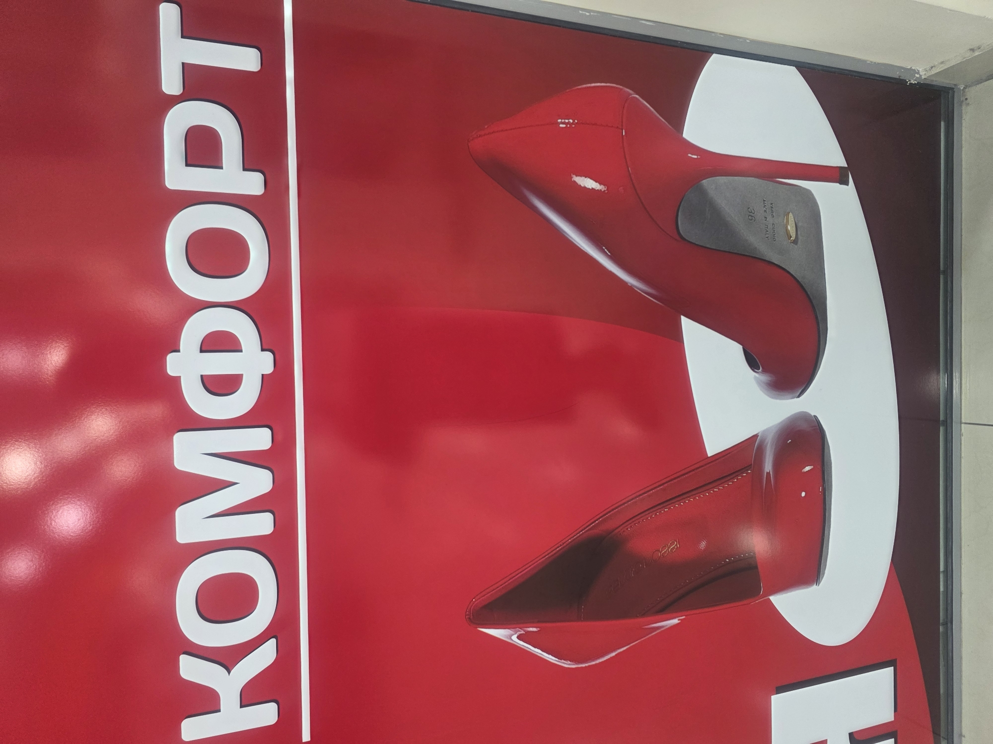 The image displays a billboard for a shoe store featuring a large, red background with white text that reads "КОМФОРТ" (Comfort). The word is centered at the top of the billboard. Below the text, two red high-heeled shoes are prominently displayed on white oval platforms. The left shoe is shown from the side, revealing its pointed toe and elegant silhouette. The right shoe is angled to show its heel and sole, which is marked with "VERO CUOIO MADE IN ITALY" and the size "36". The billboard appears to be an advertisement for a shoe store, likely located in Bila Tserkva, Ukraine, given the context of the query. The overall impression is one of luxury and style.