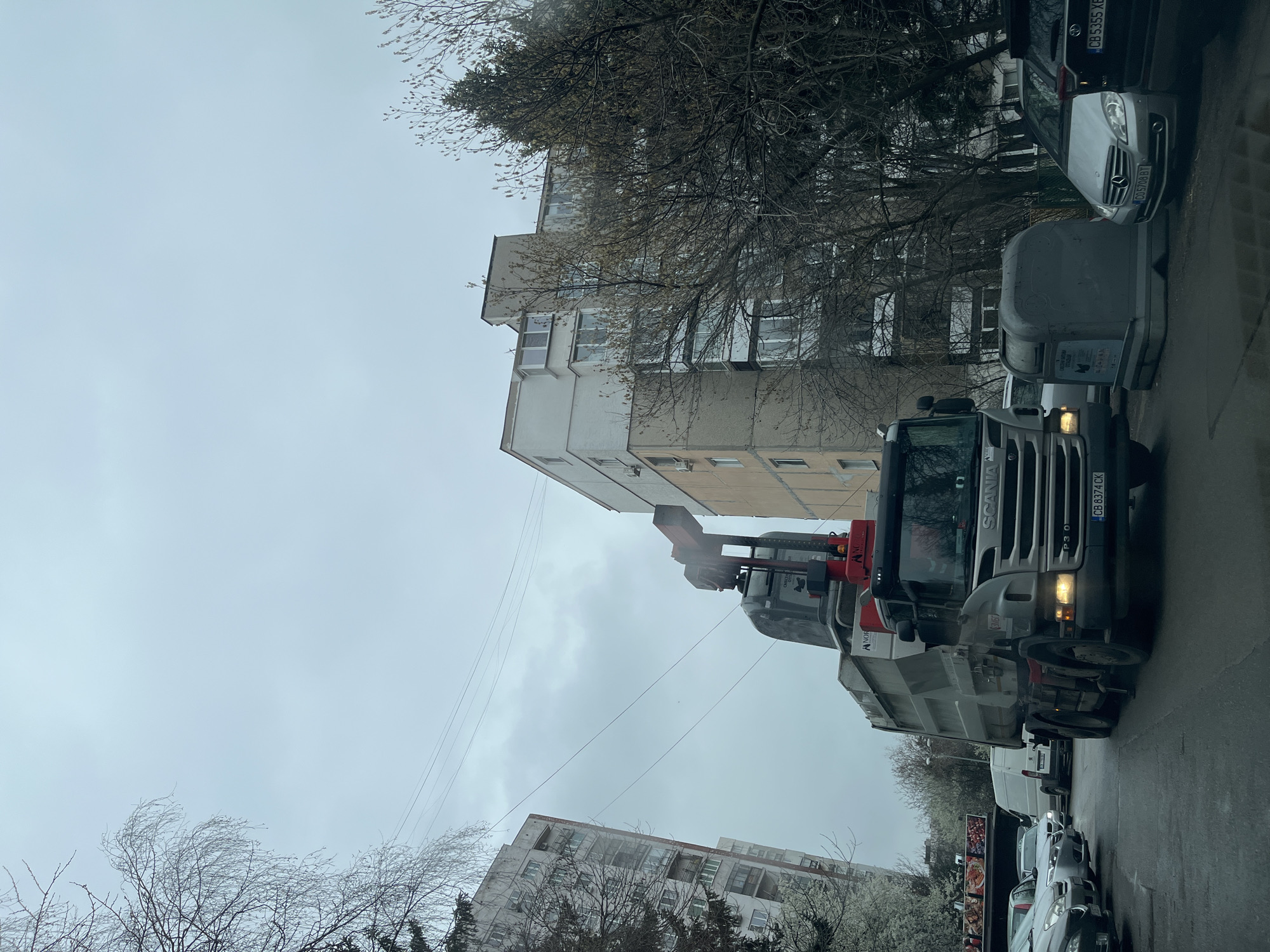 A garbage truck is parked on a city street in Sofia, Bulgaria. The truck is a Scania P310, with license plate CB 8374 CK. The truck's mechanical arm is extended over a large, grey garbage container. Behind the garbage truck are several parked cars, including a silver Mercedes-Benz with license plate CB 5709 BT and a black car with license plate CB 5355 XB. Buildings, including a multi-story apartment building, are visible in the background behind bare trees. The sky is overcast and grey.