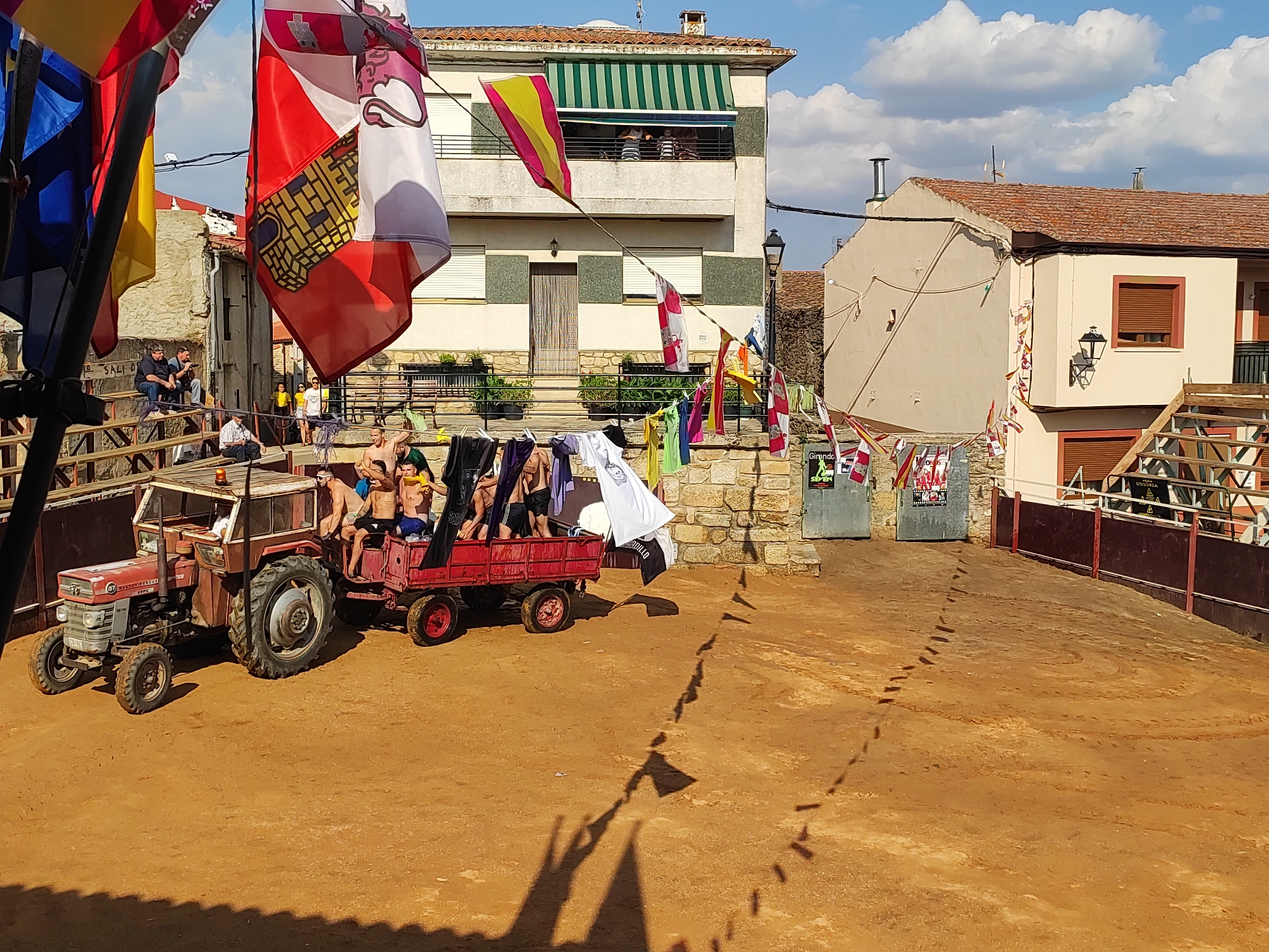 This image captures a lively outdoor scene in the city of Sobradillo, Spain. In the foreground, a vintage red tractor, a Massey Ferguson model, is parked on dusty, orange-colored ground, likely a bullring or arena. A red trailer is hitched to the tractor, carrying a group of young men, some shirtless and others in dark shorts, who appear to be celebrating or participating in an event. They are holding onto the sides of the trailer and some are waving their arms.
Dominating the background are buildings with terracotta tiled roofs and cream-colored facades. One building features a green and white striped awning, and several windows are visible. Flags of various designs and colors are prominently displayed throughout the scene, attached to poles and strung across the area. Some flags are large and unfurled, while others are smaller and fluttering in the breeze. Laundry hangs on a clothesline between two buildings, adding a touch of domesticity to the festive atmosphere.
The ground is marked with tire tracks and what appear to be footprints or hoof prints. The sky is a vibrant blue with scattered white clouds. The overall impression is one of a community gathering, possibly a festival or celebration, taking place in a traditional Spanish setting. The presence of what looks like a bullring suggests the event might be related to a local fiesta involving bulls.