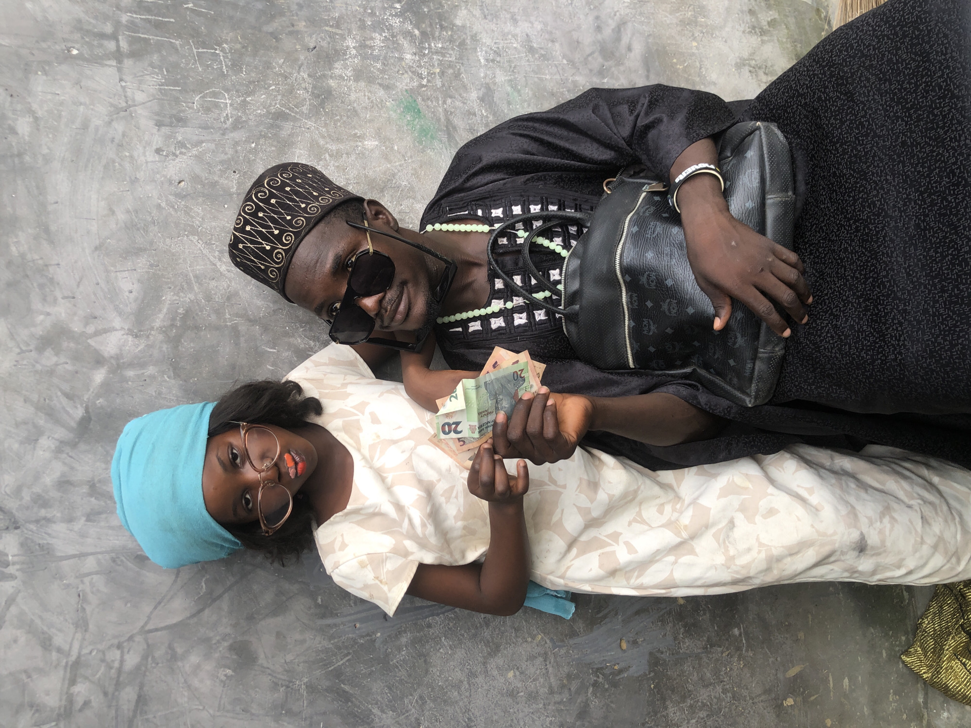 A young girl, dressed in a white patterned dress and a turquoise headscarf, poses with a man wearing a black traditional outfit and a patterned cap. The girl is wearing large, round glasses and has bright orange and red lipstick on. She holds several Nigerian Naira banknotes in her hands. The man is wearing sunglasses and a green beaded necklace. He has a black bag with a repeating "MCM" logo on his lap. They are both leaning against a textured, grey concrete wall. The scene appears to be outdoors, possibly in a street or courtyard in Ado, Nigeria. A portion of a gold-colored fabric is visible in the lower left corner of the image.