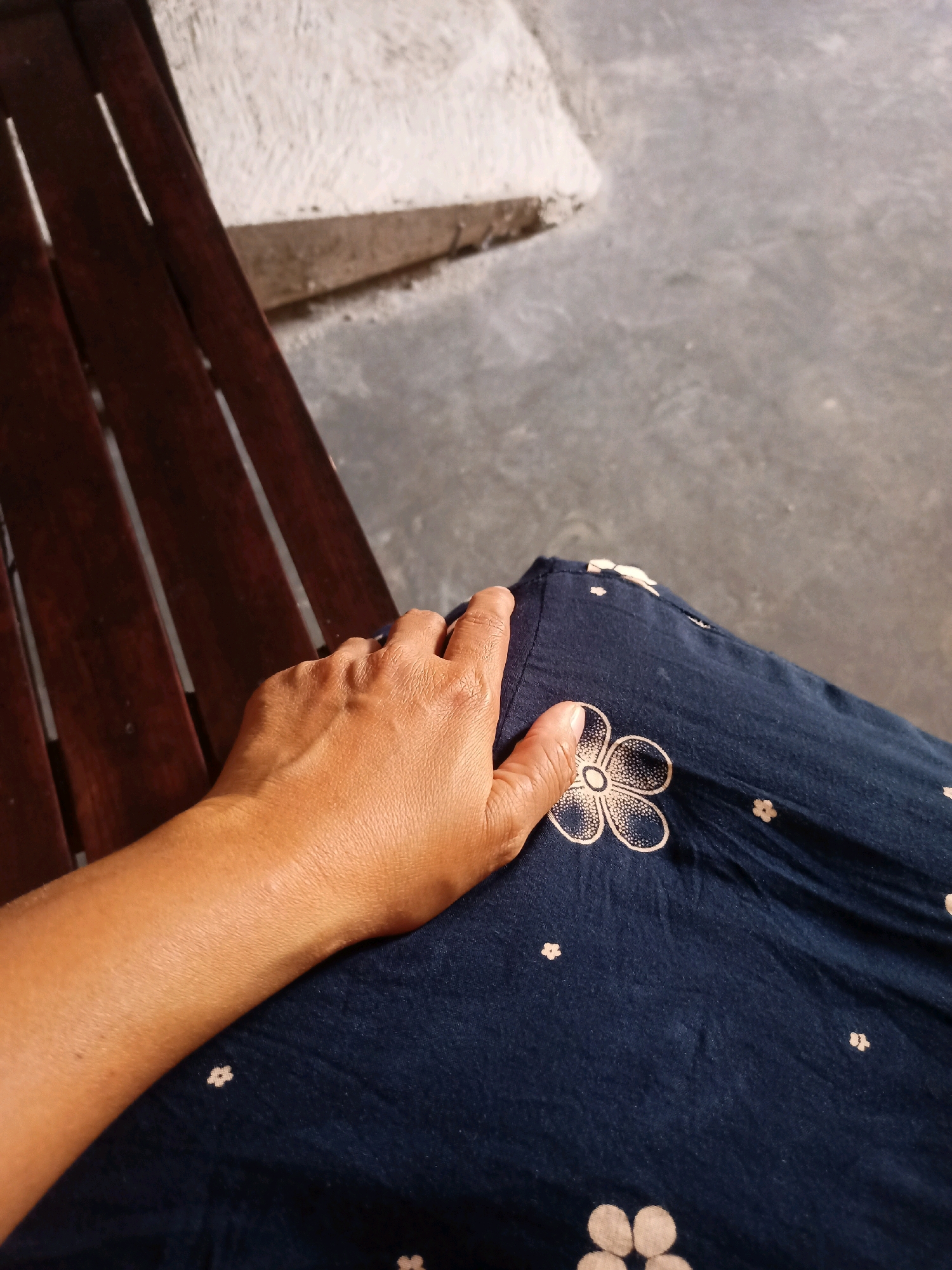 A close-up, overhead shot shows a person's hand resting on a dark blue fabric with a floral pattern. The fabric appears to be part of clothing, possibly a skirt or dress, as it drapes over what looks like a wooden bench. The bench is visible on the left side of the image, with its dark, slatted surface. The background consists of a grey concrete floor and a lighter-colored textured surface, possibly a wall or step. The image is taken outdoors, likely in a public area.