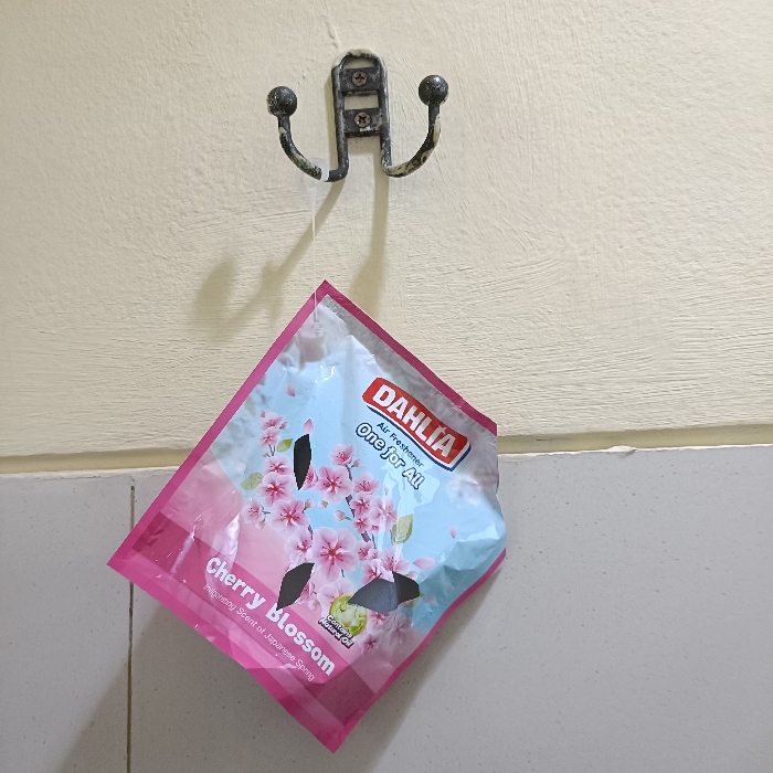 A black metal hook with two prongs is mounted on a cream-colored wall. A pink air freshener with the brand "DAHLIA" and the scent "Cherry Blossom" is hanging from the hook by a string. The air freshener has a picture of cherry blossoms on it and says "Air Freshener One for All" on it. The air freshener is hanging on a white wall. The coordinates -6.973, 110.326 place this location in Tugu, Indonesia.
