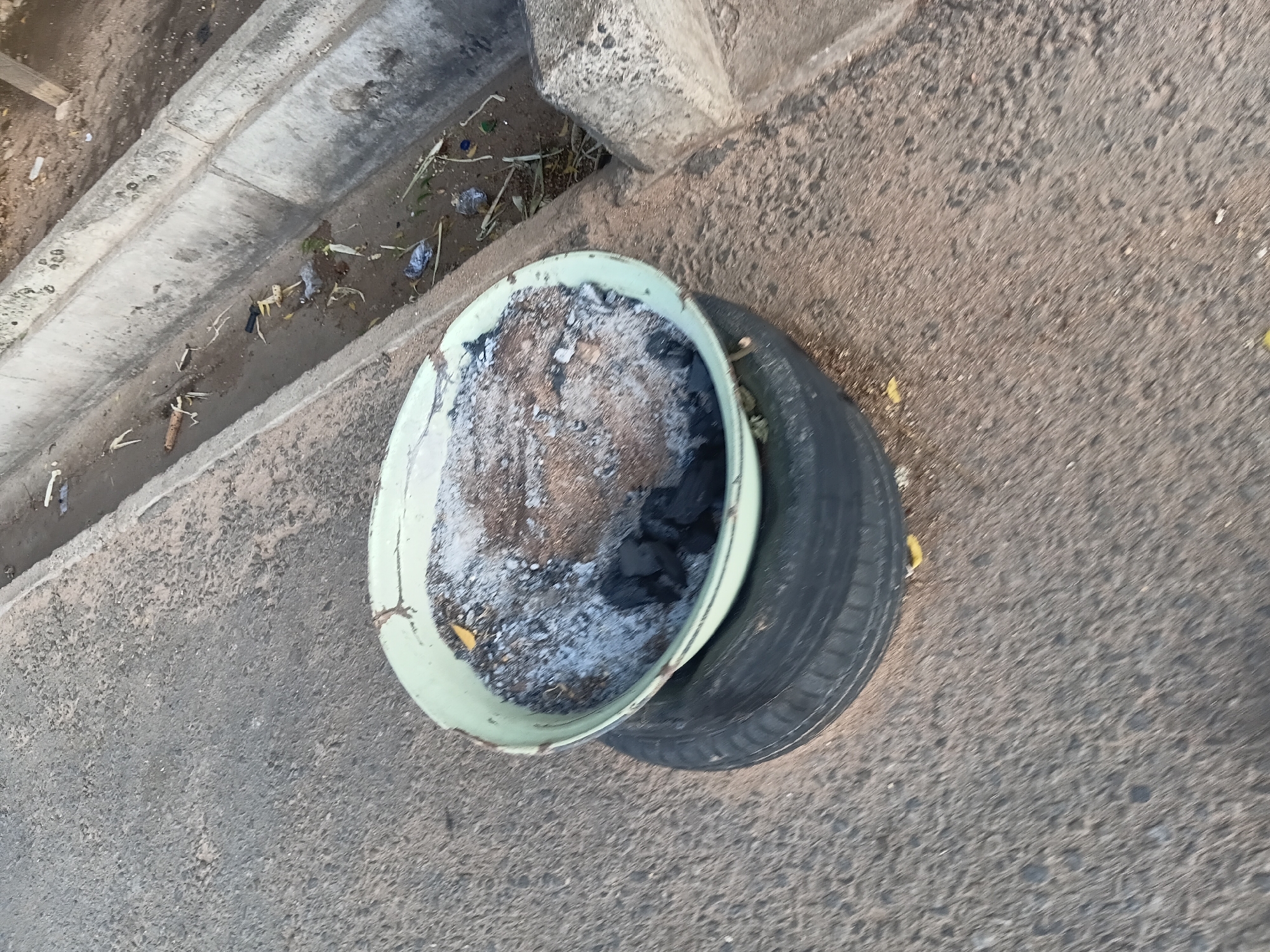 This image captures a scene on an asphalt road in Jalingo, Nigeria. In the foreground, a black tire lies flat on the ground. Resting on top of the tire is a light green metal basin. The basin is filled with ash, some charcoal, and debris, suggesting it may have been used for burning or cooking. To the right of the basin and tire, a concrete drainage ditch runs parallel to the road. The ditch contains some murky water and scattered litter, including plastic fragments and organic material. The overall environment appears to be an outdoor urban setting, possibly a roadside. There are no people visible in the image.