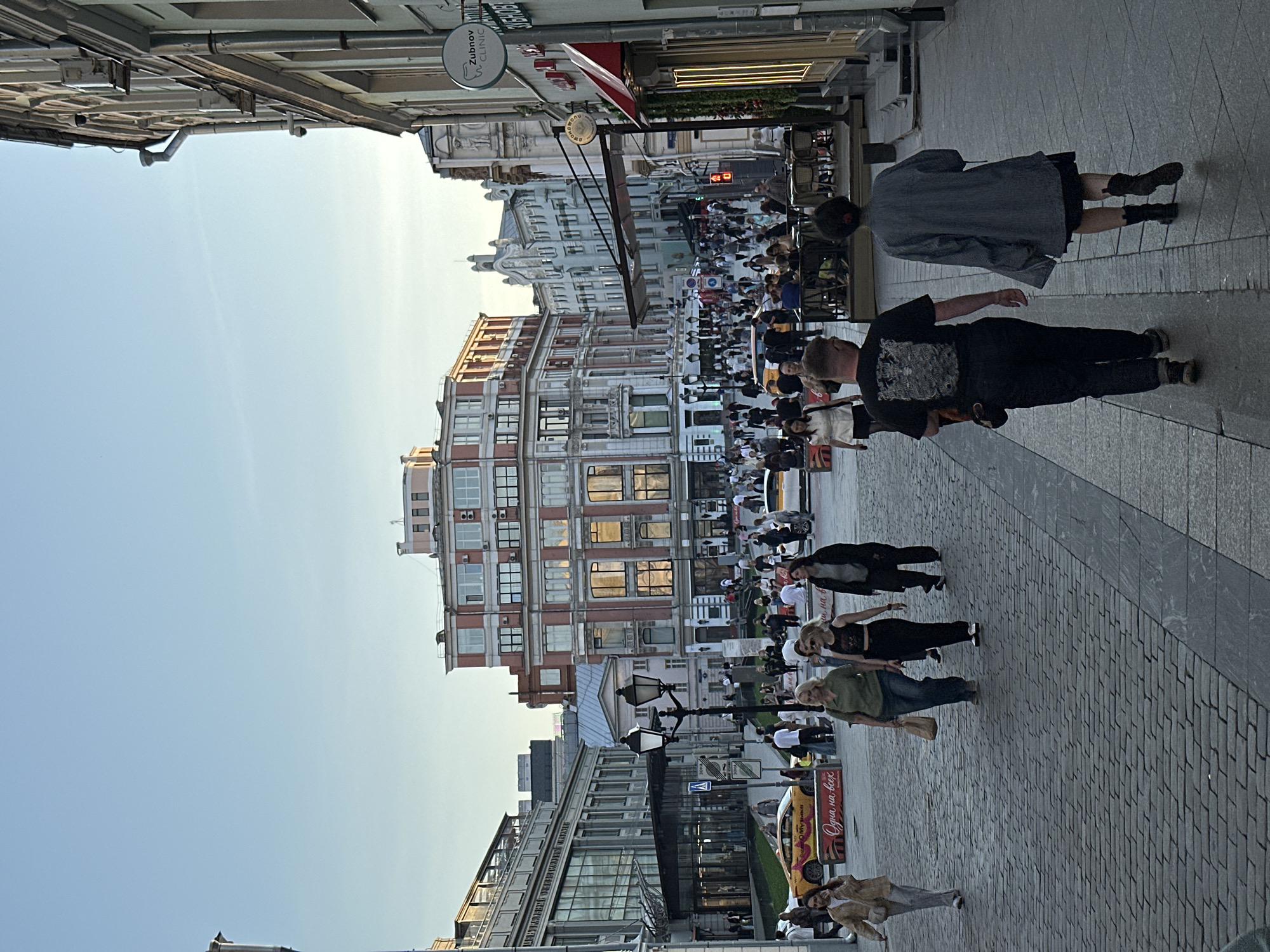 The image depicts a crowded pedestrian street in Moscow, Russia. The street is paved with cobblestones and lined with buildings of various architectural styles, including some with ornate facades and others with modern glass fronts. A large crowd of people is walking in both directions, with several individuals visible in the foreground. There are street lamps, signs, and advertisements along the street. In the upper left corner, a sign reads "Zubnov Clinic." The sky is clear and blue. The overall impression is that of a bustling city center.