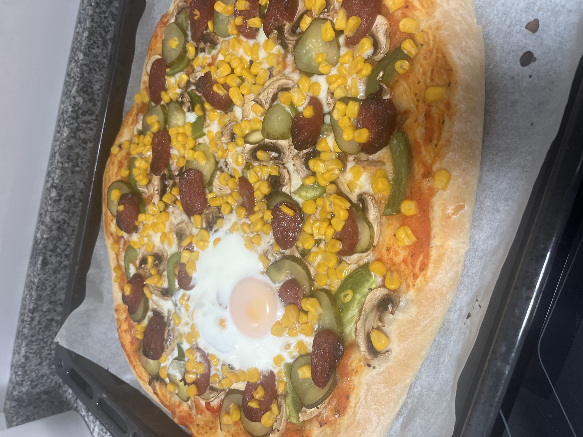 The image shows a freshly baked pizza on a baking sheet. The pizza has a thick crust and is topped with tomato sauce, cheese, corn, sausage, pickled cucumbers, mushrooms, and a fried egg in the center. The baking sheet is lined with parchment paper and is resting on what appears to be a kitchen counter. The image does not provide any information to verify the location as the city of Mogán, Spain.