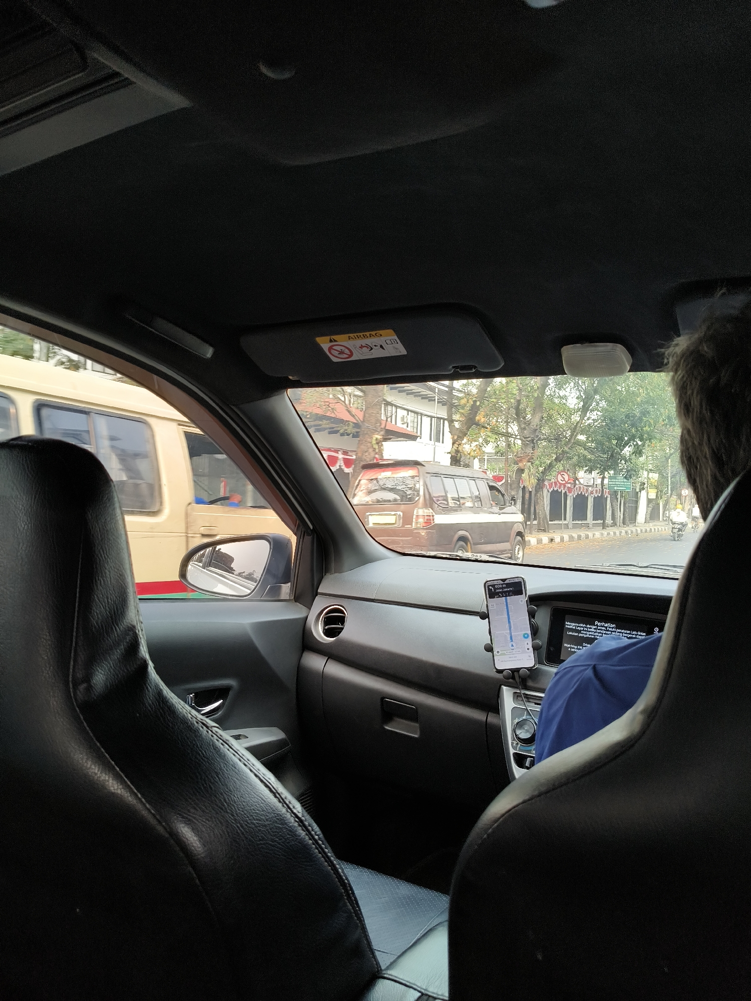 The image is a point-of-view shot from inside a car, looking towards the dashboard and out the front and side windows. The interior of the car appears to be black, with textured upholstery on the seats and dashboard. A smartphone is mounted on a dashboard holder, displaying a navigation application. The driver, partially visible from the back of their head and shoulders, is wearing a blue shirt. Outside the car, traffic is visible, including a light-colored bus to the left and a dark-colored SUV in front. The street scene includes trees, buildings, and other vehicles, suggesting an urban environment. The image appears to have been taken in Bandung City, Indonesia, based on the license plates of some vehicles and the context of other images in the dataset.