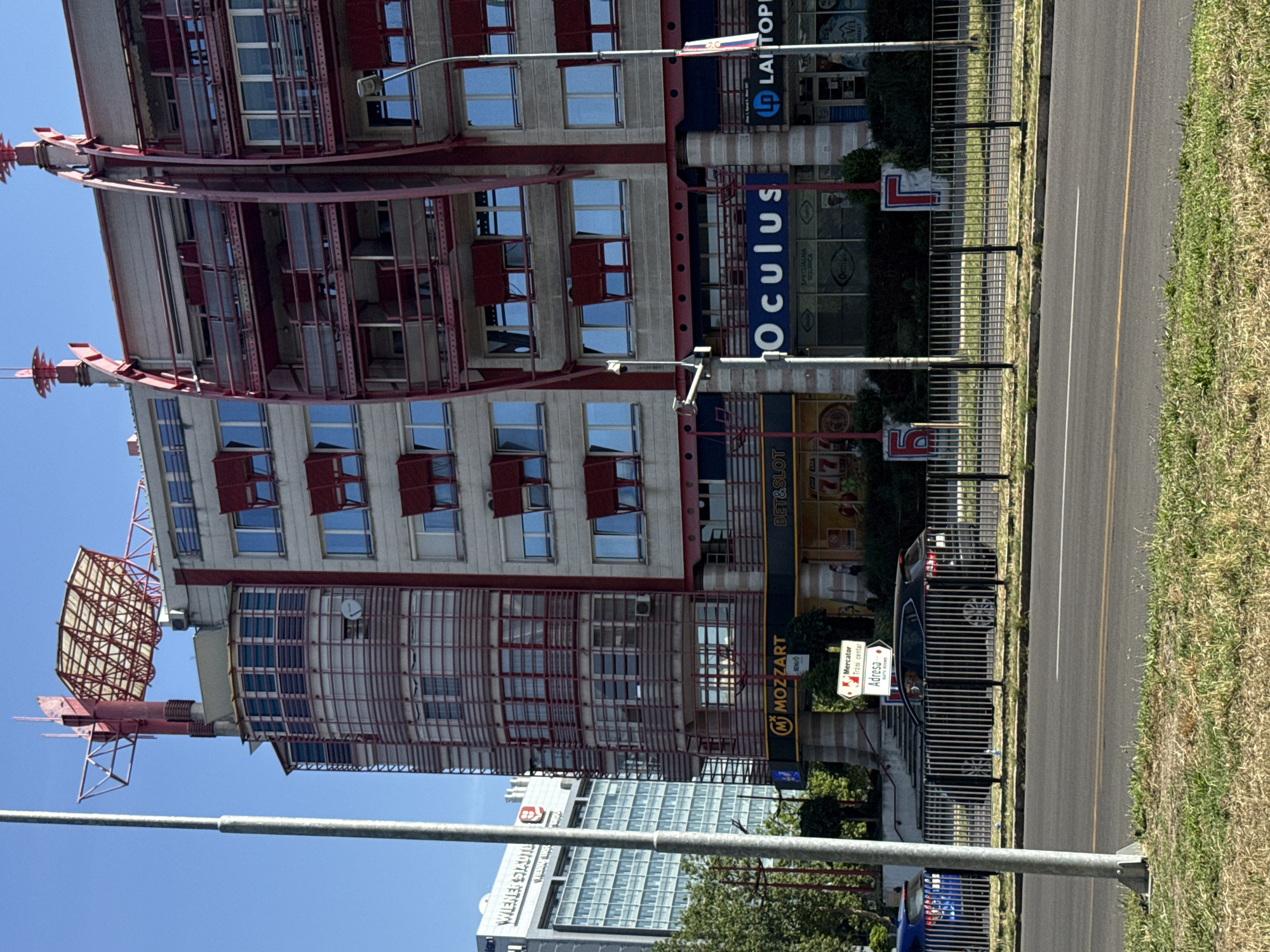 This is a photograph of a modern building in the city of Belgrade, Serbia, taken from across a street. The building features a grey facade with red architectural elements, including balconies and a prominent red structure on top. Several businesses are visible at the ground level, including "MOZZART," "BET&SLOT," "Oculus," and "LAITOP." A black car is parked in front of the building behind a black metal fence. To the left of the building, another taller, modern building with a sign that reads "WIENER STAEDTL" is partially visible. The sky is clear and blue, and the scene is illuminated by bright daylight.
