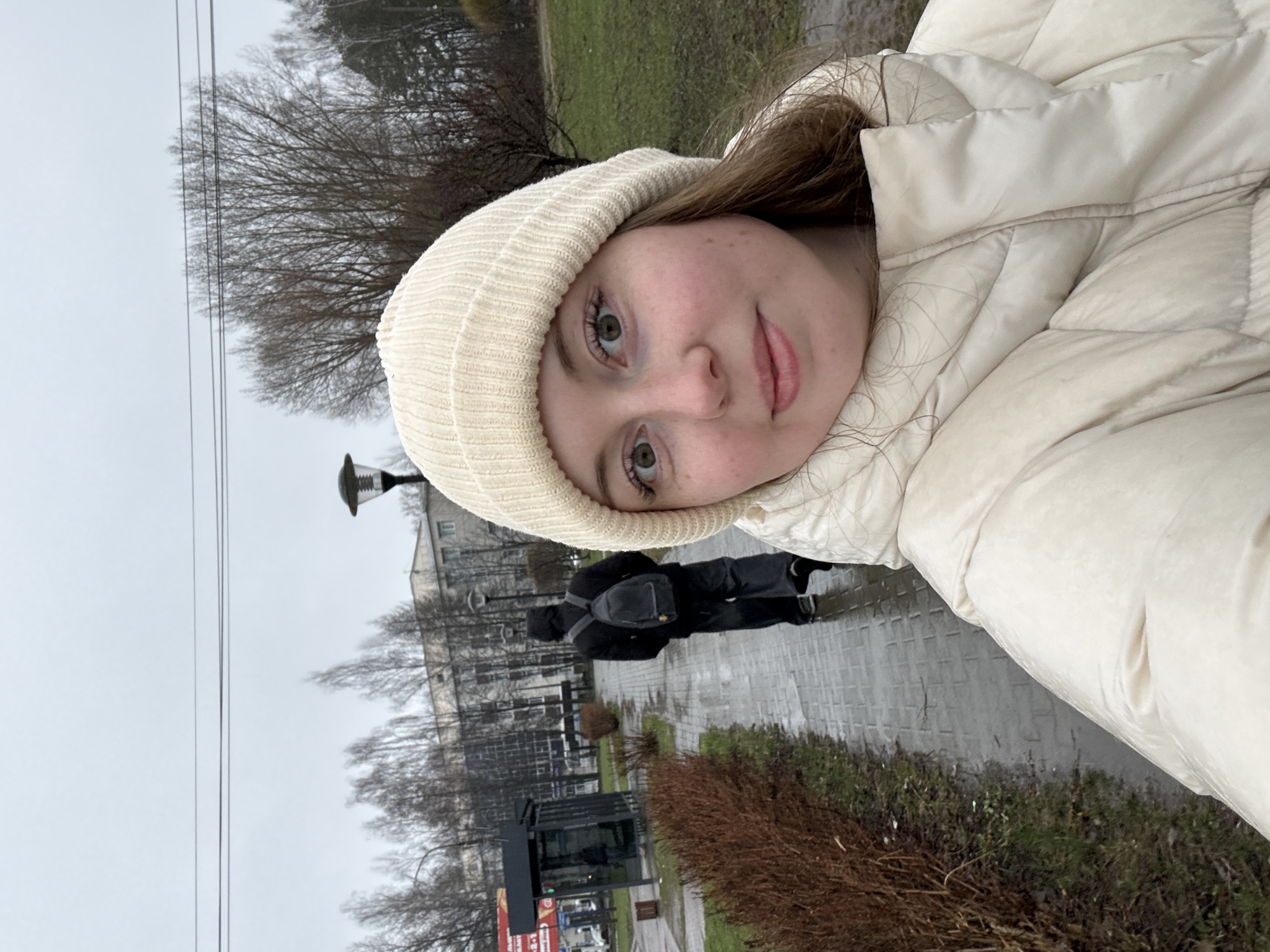 The image is a rotated selfie of a young woman outdoors on an overcast day. She has light skin, light brown hair, and green eyes, and is wearing a light-colored (beige or cream) knit beanie and a matching puffer jacket. She looks directly at the camera with a neutral expression and slightly parted lips.
The background features a grey, overcast sky with bare, deciduous trees, indicating a cooler season like late autumn, winter, or early spring. A wet, paved path or sidewalk runs through the scene, suggesting recent rain. In the mid-ground, another person, dressed in dark clothing with a backpack, walks away on the path.
Further back, there are buildings and a street light. Overhead, utility wires are visible against the sky. On the ground, brown and green shrubbery lines the path. Near a dark structure, possibly a kiosk or bus stop, a sign with visible text reads "24 ЧАСА" (24 HOURS) and "20-80%" in red, implying a commercial establishment in a region using Cyrillic script. The overall scene depicts a casual moment in a damp, urban or suburban environment.
