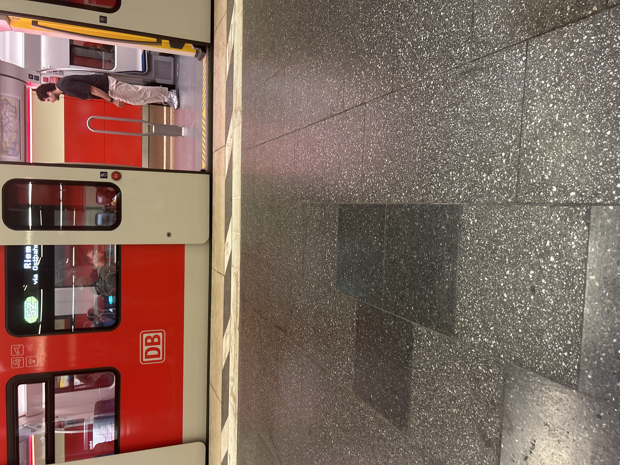 The image displays a red and white S-Bahn train, identified by the prominent "DB" (Deutsche Bahn) logo on its side, stopped at a platform. The train's doors are open, revealing its interior. Inside the train, near the open door, a person with dark, curly hair, wearing a dark t-shirt and light-colored pants, stands facing deeper into the train car. A silver metal handrail or barrier is visible inside the doorway.
The platform floor is made of large, dark grey speckled tiles. A yellow tactile paving strip runs along the edge of the platform next to the train. Reflections of lights or ambient sources are visible on the platform surface, suggesting either artificial lighting or indirect daylight.
On the train's window, a digital display shows "S2" and "Riem via Ostbahn," indicating the train line and its destination. Various accessibility and safety symbols, such as a bicycle icon, wheelchair icon, and an emergency stop button symbol, are visible near the train door. The overall scene captures a moment at a train station, likely during the daytime, with passengers potentially boarding or disembarking.
