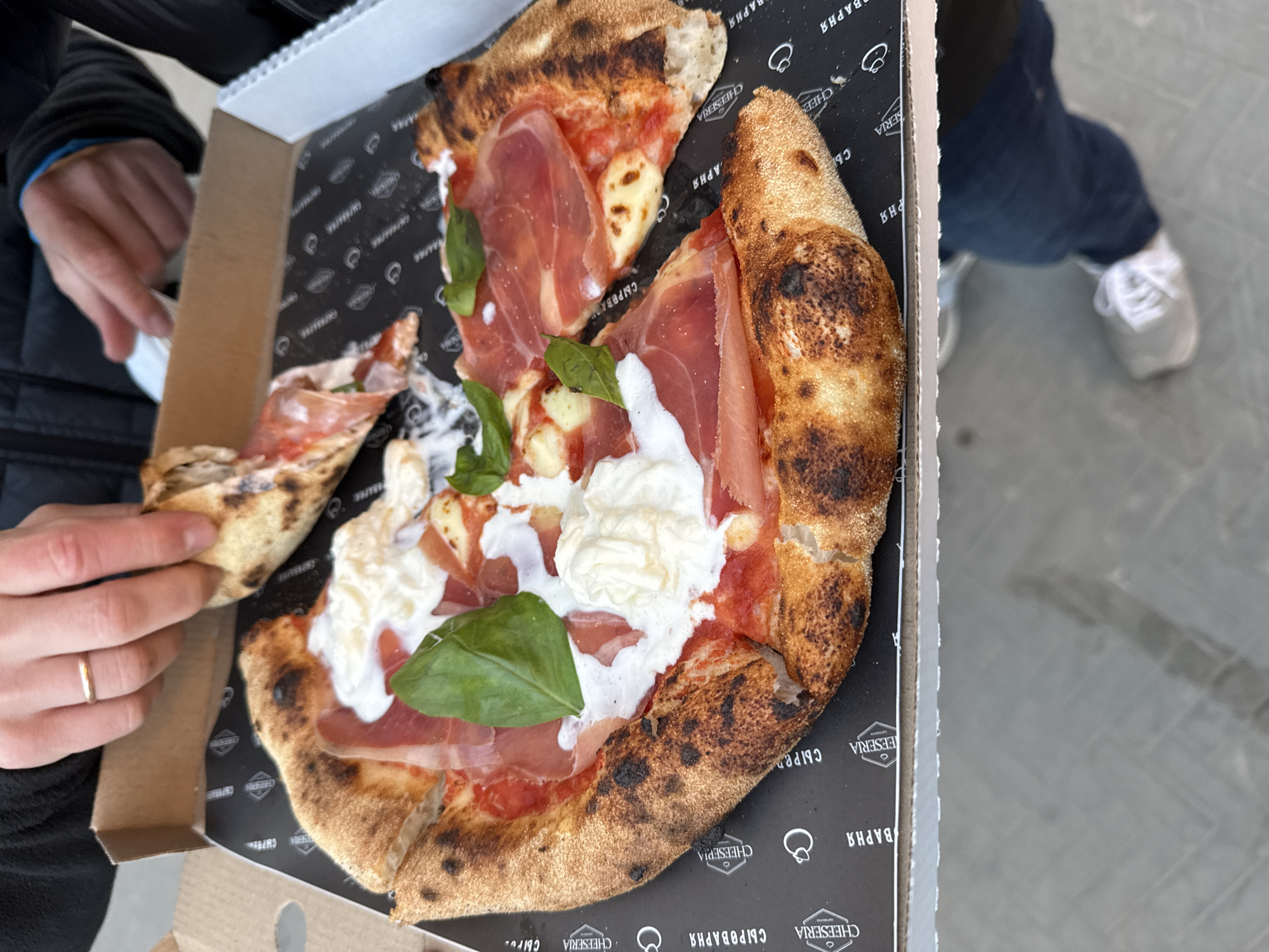 A high-angle shot shows a partially consumed pizza inside its delivery box. The pizza, which has a charred crust, is topped with tomato sauce, prosciutto, mozzarella, and basil leaves. A hand, wearing a gold band on the ring finger, holds a slice of the pizza. The pizza box has a black interior printed with the words "CHEESERIA" and "СЫРОВАРНЯ," which translates to "Cheese Dairy." These repeated words, along with a logo, indicate the branding of the establishment. Another leg with a white sneakers is visible in the background. The environment is a light grey pavement. The pizza's charred crust and unique toppings suggest a gourmet or artisanal pizza offering. Given the name "CHEESERIA" and its Russian equivalent, the pizza's origin is likely in Russia, specifically in Kazan, Russia.
