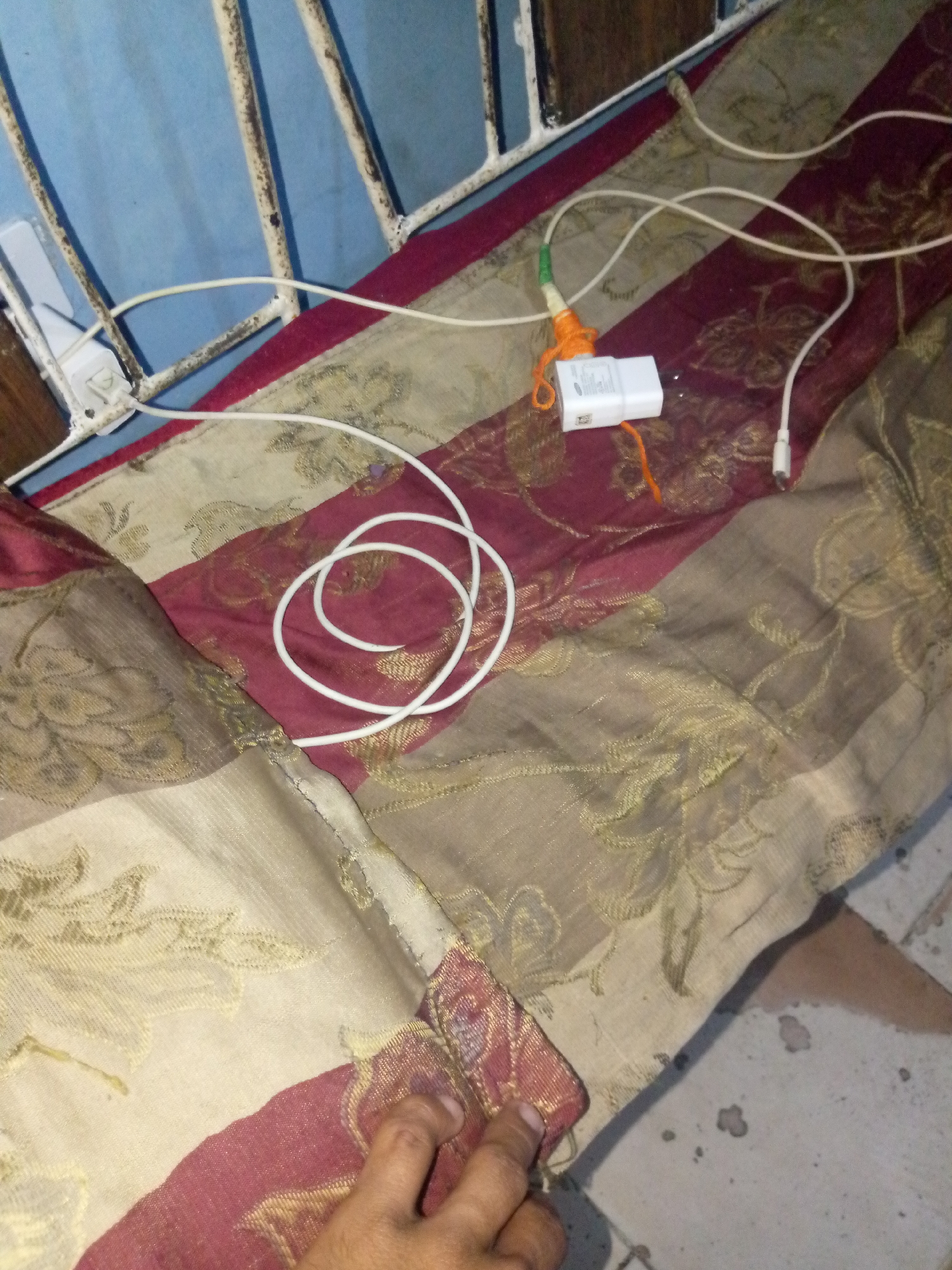 A white phone charger and its white cable are on top of a colorful patterned fabric. The fabric has stripes of dark red, light beige, and gold. The charger is plugged into a white power adapter that is connected to a white cord. The cable is coiled and draped across the fabric. A person's hand is visible at the bottom right of the image, touching the fabric. The background appears to be a blue wall with a white metal railing. The scene is indoors, possibly in a bedroom or living area.