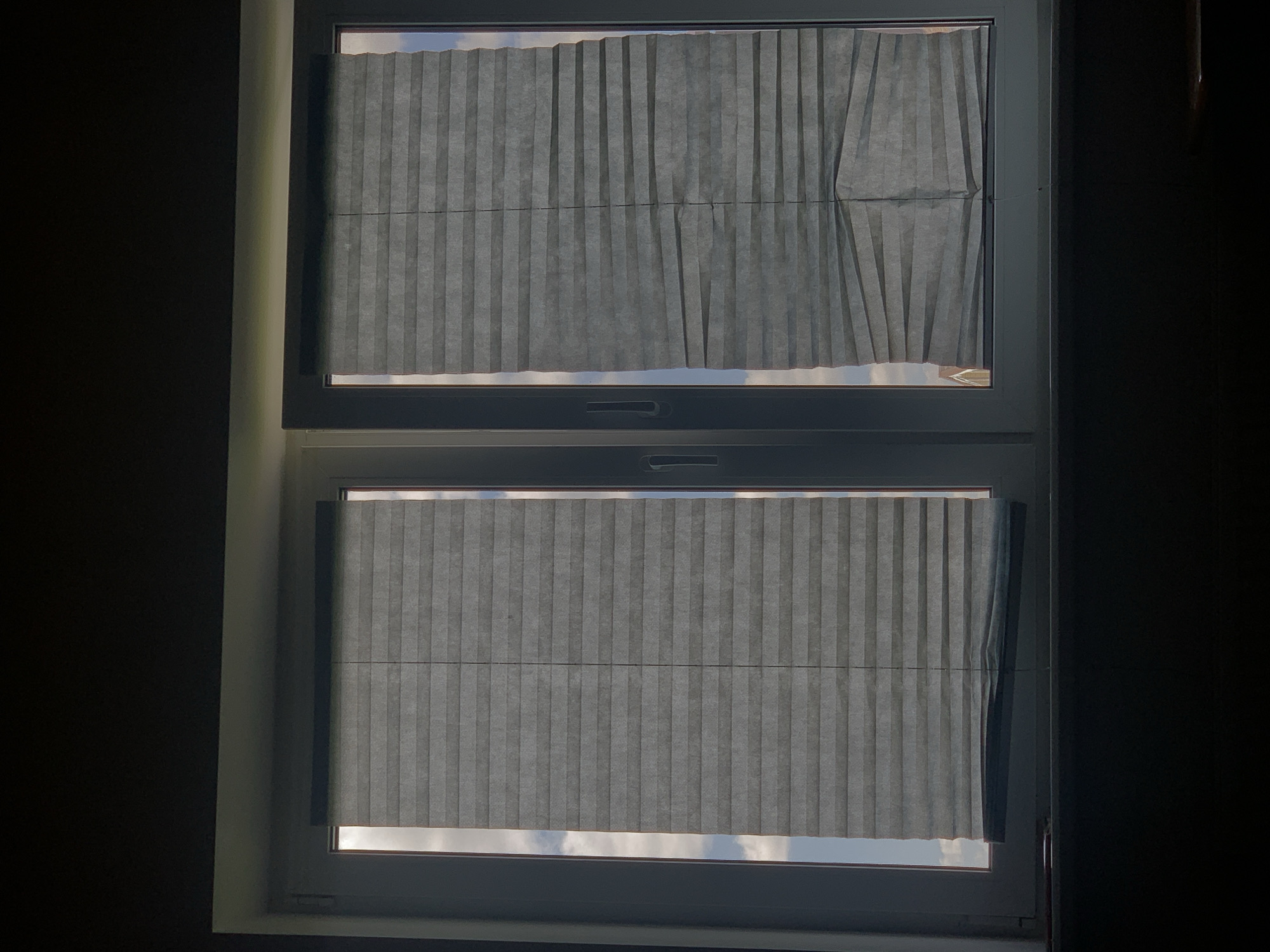 The image shows a close-up view of a window with two sets of pleated blinds. The blinds are gray and appear to be partially drawn, allowing some light to filter through and revealing a cloudy sky and what appears to be a building with a red roof in the distance. The window frame is white, and there are handles on the window panes. The lighting suggests it is daytime, but the blinds are obscuring a clear view of the outside. The scene is devoid of any people or any indication of activity. The location of the image is not specified, and there is no information within the image itself that would suggest it is in Minsk, Belarus.