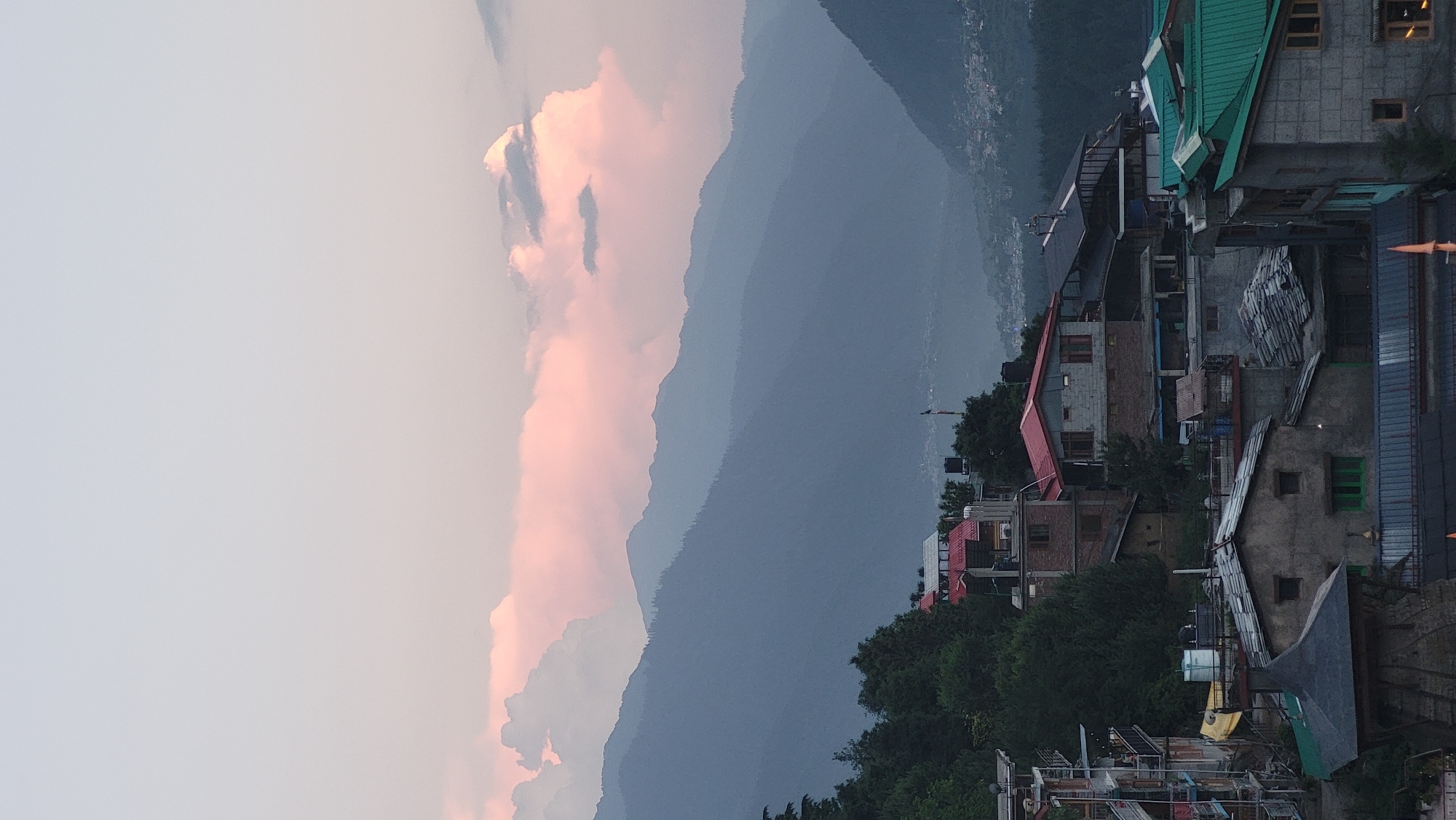The image captures a serene twilight scene in Vashisht, India, showcasing a mountainous landscape dotted with buildings. The setting is primarily natural, featuring majestic mountains that stretch across the midground, displaying layers of blue and grey tones as they recede into the distance, indicative of atmospheric perspective. A distant valley, visible between the mountain ranges, shows faint urban lights, suggesting a settlement below.
The sky above is a canvas of soft hues, dominated by a large, prominent cloud on the left side that glows with delicate pink and orange tones, suggesting either a sunrise or sunset. The rest of the sky is a lighter, muted grey-blue.
In the foreground and mid-right, a collection of buildings is visible, characteristic of a mountain village or town. These structures vary in design, with some featuring bright red or green sloped roofs, while others have flatter grey or metallic roofs. The buildings appear to be residential or guesthouses, nestled among lush green trees, which punctuate the built environment. No people are visible, and no specific activity is taking place, contributing to the tranquil atmosphere. The overall lighting is dim, consistent with early morning or late evening. There is no visible text in the image.