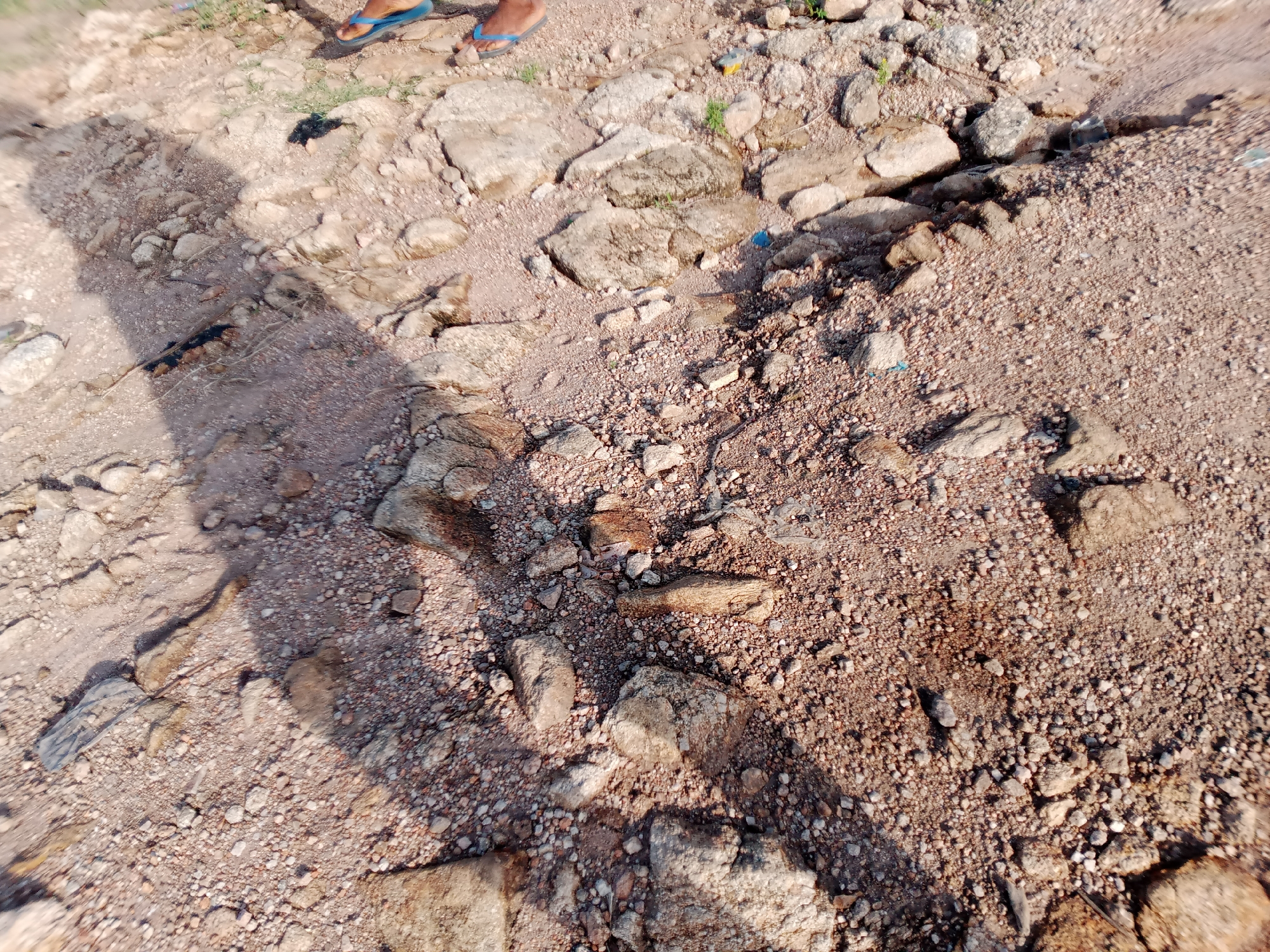 The image depicts a rocky and sandy terrain, likely an outdoor area in Jalingo, Nigeria. In the upper portion of the image, the feet of at least two individuals wearing blue flip-flops are visible. The ground is covered with a mixture of coarse sand, gravel, and various sized rocks. Some sparse green vegetation is also present amongst the rocks. A prominent shadow stretches across the left side of the frame, suggesting the presence of an unseen object or person casting it. The scene appears to be a natural or undeveloped area, possibly a pathway or the edge of a body of water, given the texture of the ground. There are a few small pieces of debris, including what appears to be plastic, scattered on the ground.