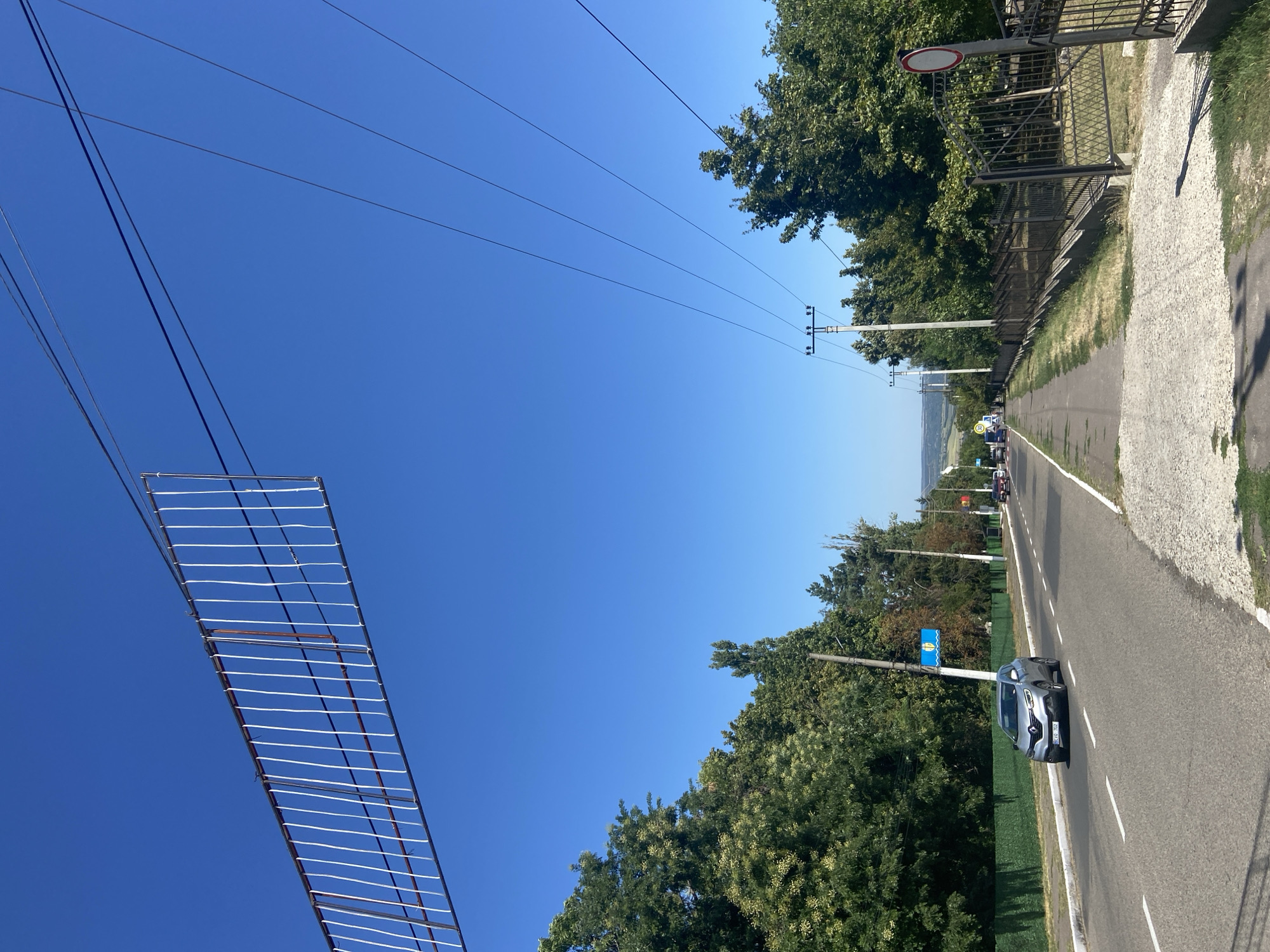 This is a full, eye-level, outdoor, daytime shot of a road in a suburban or rural area, likely in Eastern Europe given the license plate and architecture. The sky is a clear, vibrant blue. In the foreground, a paved road runs from left to right, with a white solid line marking the edge of the lane and a dashed white line dividing lanes. A silver Renault Kadjar, license plate "BEVO 702", is driving on the road, heading away from the viewer. To the left of the road is a sidewalk, and beyond that, a row of lush green trees. To the right of the road is a black metal fence, beyond which are more trees. There are several power lines crisscrossing the sky above the road. Further down the road, several other vehicles are visible, and some buildings can be seen in the distance. A blue sign with a yellow wave symbol and a white fish is mounted on a pole near the road.