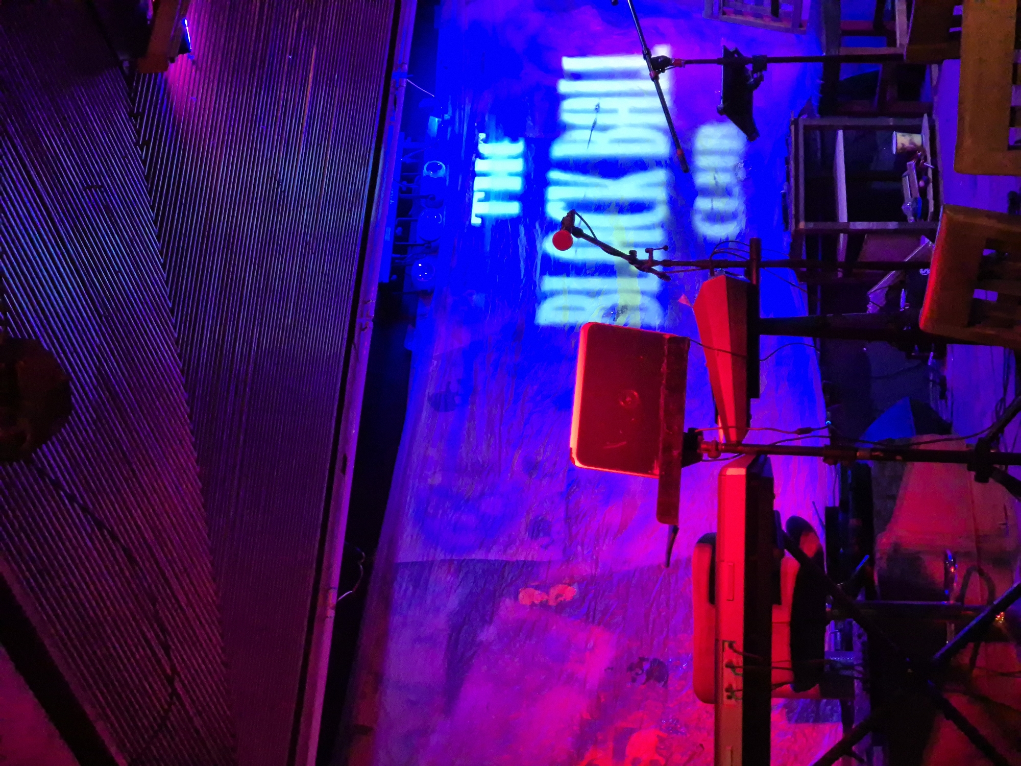 The image captures a vibrant, dimly lit interior, possibly a performance space or club. The words "THE BLACK BEAT CLUB" are projected in bright blue light against a textured wall in the background. In the foreground, there are sound equipment, including red speakers and microphone stands, situated amidst a complex arrangement of metal scaffolding. The scene is bathed in purple and red light, giving it a distinctly energetic and dynamic atmosphere. Given the club's name, the location is likely a live music venue in the city of Puebla, Mexico.