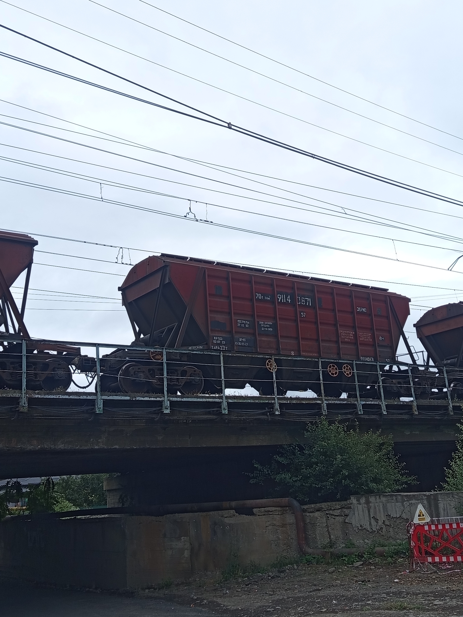 The image depicts a view from under a railway bridge in Khashuri, Georgia. A train with several open-top freight cars, painted red, is visible crossing the bridge. The cars have markings and numbers on their sides. The bridge structure is made of concrete and appears aged. Below the bridge, there is rough terrain with some overgrown vegetation and concrete structures. Overhead power lines are visible against a cloudy sky. There are no people or animals in the frame.