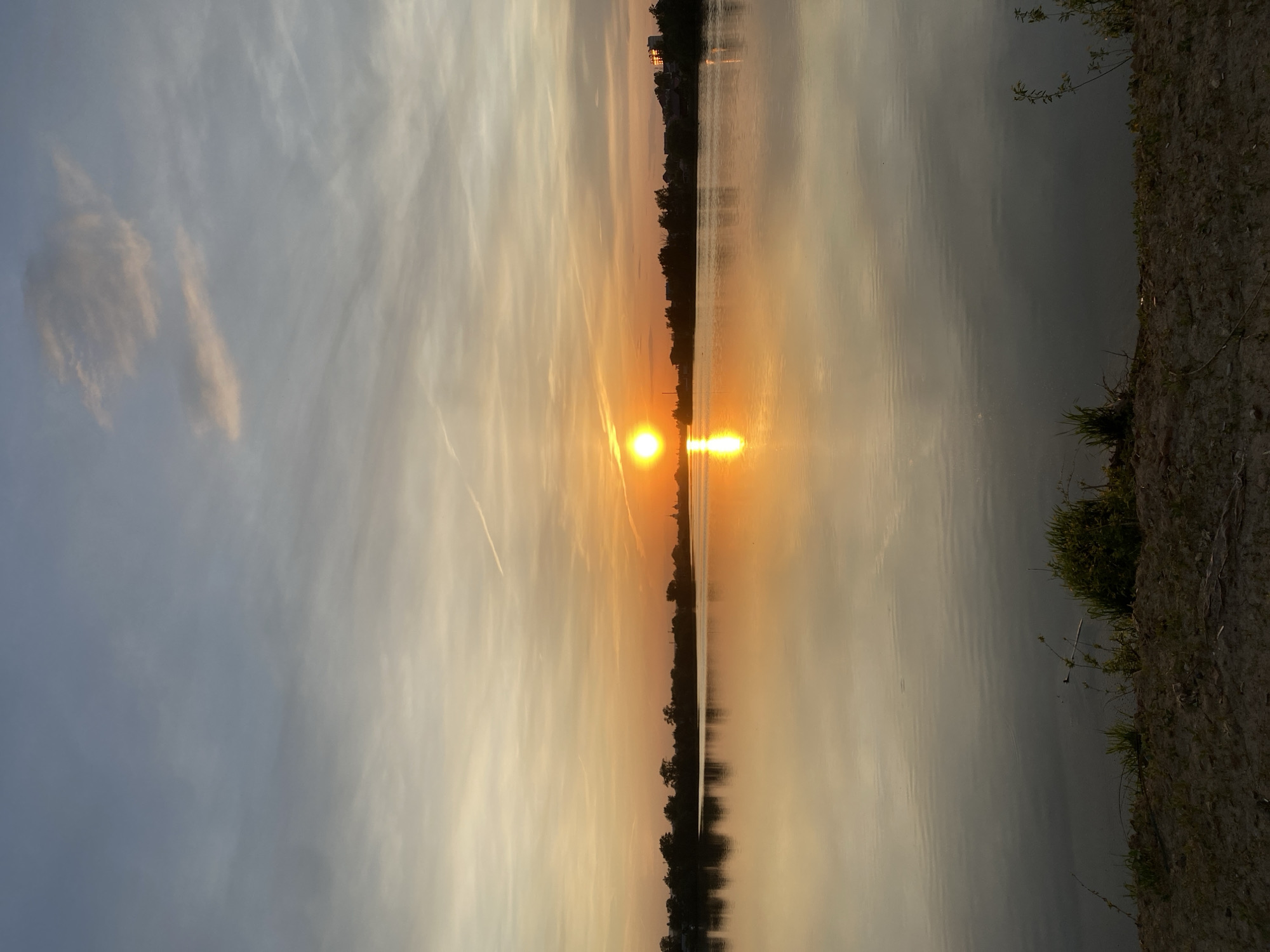 The image depicts a sunset over a body of water, likely a lake or wide river. The sun is centered, with its reflection extending vertically downward in the water. The sky is a mix of pale blue and orange, with wispy clouds streaking horizontally. The water is calm, reflecting the colors of the sky. Two strips of land, likely shorelines, run along the left and right sides of the water, with sparse vegetation visible. There are buildings in the distance. Based on the location requested, it is possible the scene is near Aramil, Russia, but there is nothing within the image itself to confirm the exact location. There are no people, actions, or interactions visible in this still scene.