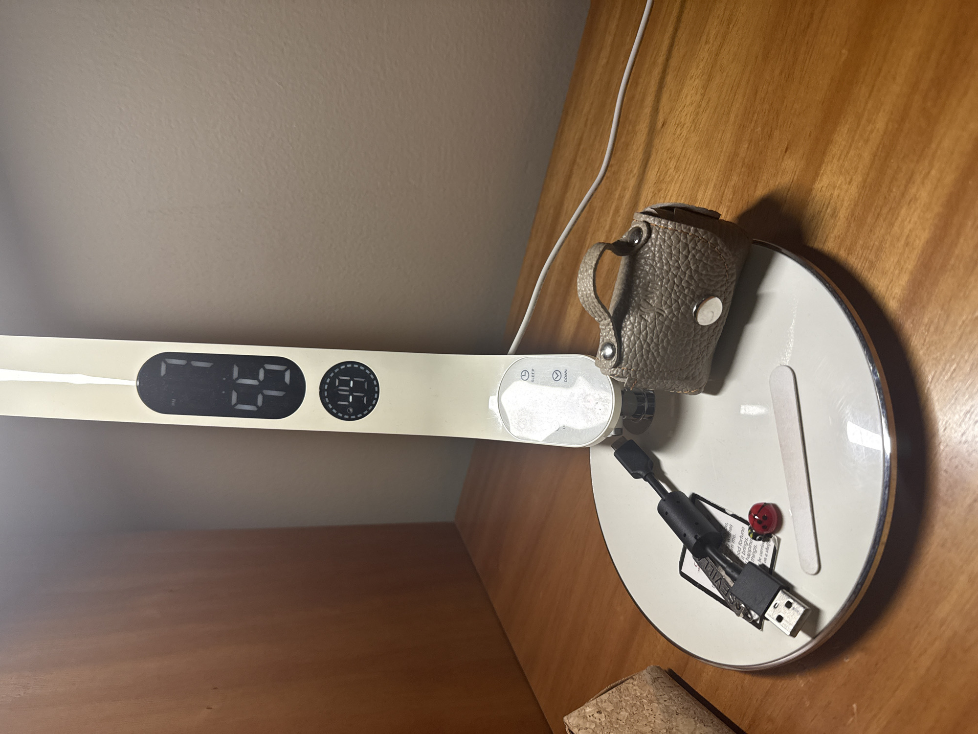 An overhead shot displays a modern desk lamp on a wooden surface, set against a light-colored wall. The scene is an indoor setting, likely in San Bernardino Tlaxcalancingo, Mexico, during the evening as indicated by the digital clock.
The lamp itself has a long, flat head with a digital display showing "7:55 PM" and a temperature of "29." Nearby buttons are labeled "SLEEP" and "DOWN." The lamp's base is round and white with a silver rim, serving as a platform for several small items. On the base, there is a small, textured, light brown pouch, possibly leather, with a handle and silver snap closures. Adjacent to it is a plain white flat stick, resembling a popsicle stick. A small, red ladybug ornament is also visible. A black USB multi-charging cable, featuring a USB-A connector and other smaller ports, is placed on the base.
Underneath the USB cable, a small piece of paper or tag contains partially visible text. Readable phrases include "wishes," "m me.", "bod fortune" (likely "good fortune"), "it brings,", "happines" (likely "happiness"), "things.", "be carrie" (likely "be carried"), and "on a shell." A white power cable runs from the lamp across the wooden surface, and a cork block or coaster is partially visible in the lower left corner. The environment appears functional and personal.