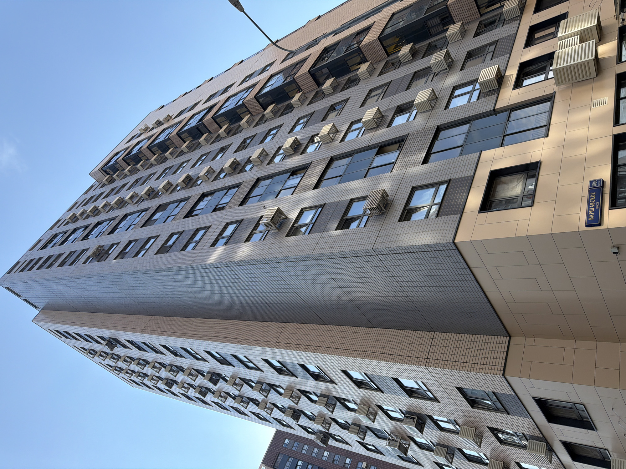 An upward, wide-angle shot captures a modern, multi-story apartment building in Moscow, Russia, against a clear blue sky. The building's facade is a combination of light grey textured tiles and beige panels, with numerous dark-framed windows and external air conditioning units mounted beneath many of them. The perspective emphasizes the building's height and the regularity of its design. A street sign with Cyrillic lettering, "ВАРШАВСКОЕ ШОССЕ 170 E," is visible on the lower portion of the building, indicating the location as Warsaw Highway 170 E. The scene is static, with no people or ongoing actions depicted. The lighting suggests it is daytime.