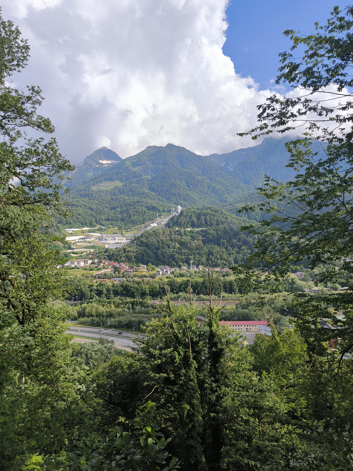 This image captures a panoramic view of Krasnaya Polyana, Russia, a mountain resort town. The foreground is dominated by lush green foliage from trees, framing the expansive landscape beyond. In the mid-ground, a town or settlement is visible, with buildings of varying sizes and architectural styles, nestled amongst verdant hills. A multi-lane highway with a few vehicles can be seen winding through the valley, indicating connectivity and infrastructure. Further in the distance, majestic mountains rise, their slopes covered in dense forests. Some of the higher peaks show patches of snow, suggesting a significant altitude. The sky is a mix of bright blue patches and dramatic, fluffy white clouds, hinting at a dynamic weather system. The overall scene depicts a harmonious blend of natural beauty and human development in a mountainous setting.