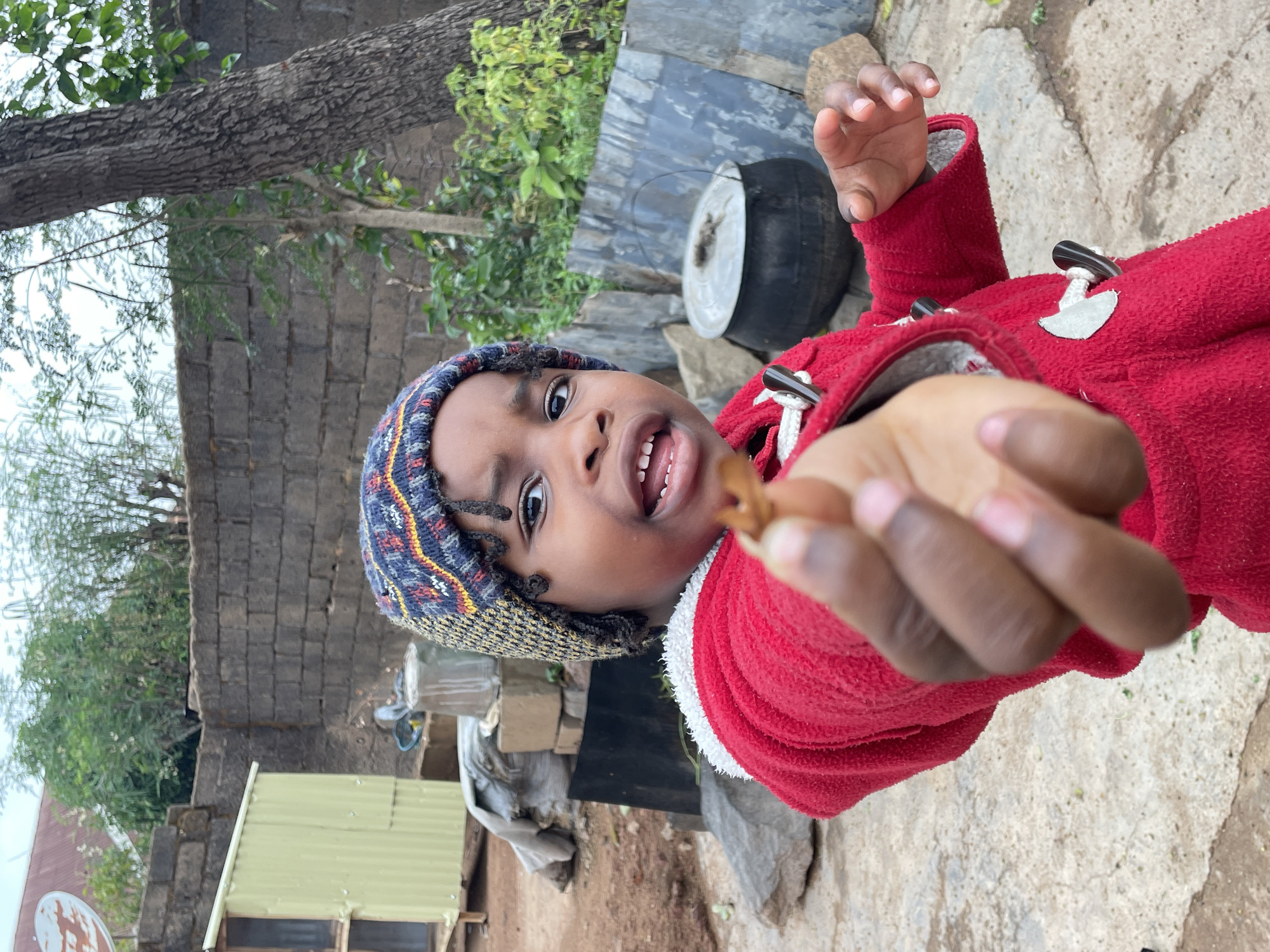 The image depicts a young child wearing a blue and multicolored knit hat and a red hooded coat standing in an outdoor setting. The child is holding out a small brown object, possibly food, towards the viewer. The background includes a brick wall, green foliage, a metal container, and what appears to be a cooking pot. There is also a corrugated metal structure visible. The scene is likely in a residential area in Jos, Nigeria.