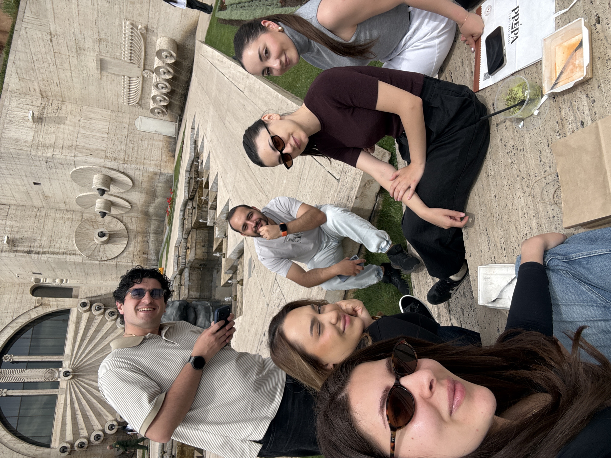 The image shows a group of six people sitting on a stone surface, likely at the Cascade in Yerevan, Armenia. The Cascade is a large, monumental stairway with fountains and sculptures. The background features architectural elements of the Cascade.
The people are positioned in a somewhat circular arrangement, suggesting a casual gathering. They appear to be eating or having a meal, with containers and bags of food visible. Some are looking at the camera, while others are engaged in conversation or looking at their phones. Their clothing styles are casual and contemporary. The light appears to be natural daylight.