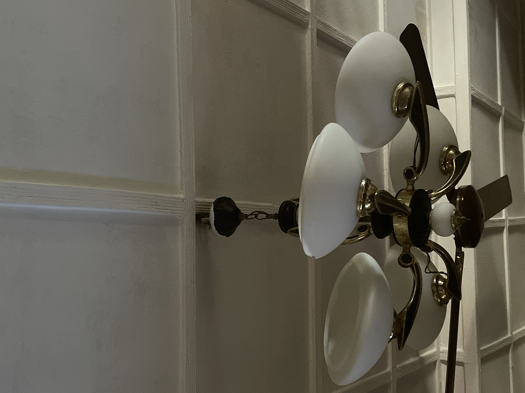 A ceiling fan with a chandelier attachment is shown. The chandelier has a dark central body with brass accents and five white glass shades. Three shades are visible from this angle, each appearing to be an oval shape. The ceiling fan blades are dark brown and are positioned beneath the chandelier. The ceiling is white and features a coffered design. The image was taken in Kakau, Nigeria.