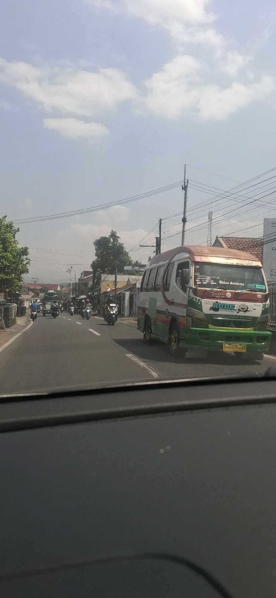 The image captures a street scene in Sukasari, Indonesia, on a bright, cloudy day. A white and red passenger van, adorned with various decals including "RUDEBOX" and "Bukan Selebrms," is driving on the right side of the road. In front of the van, there are several motorcycles on the road. On the left side of the road, a green truck is visible, also with motorcycles. The environment consists of low-rise buildings, trees, and utility poles with visible overhead wires. The foreground shows the interior of a vehicle, suggesting the photo was taken from a passenger's perspective.
